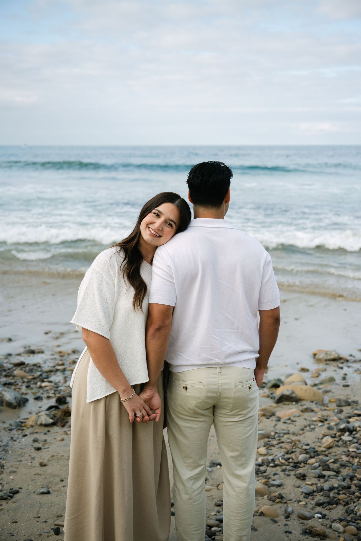 Smiling woman leans on mans shoulder as couple holds hands facing ocean on rocky beach.
