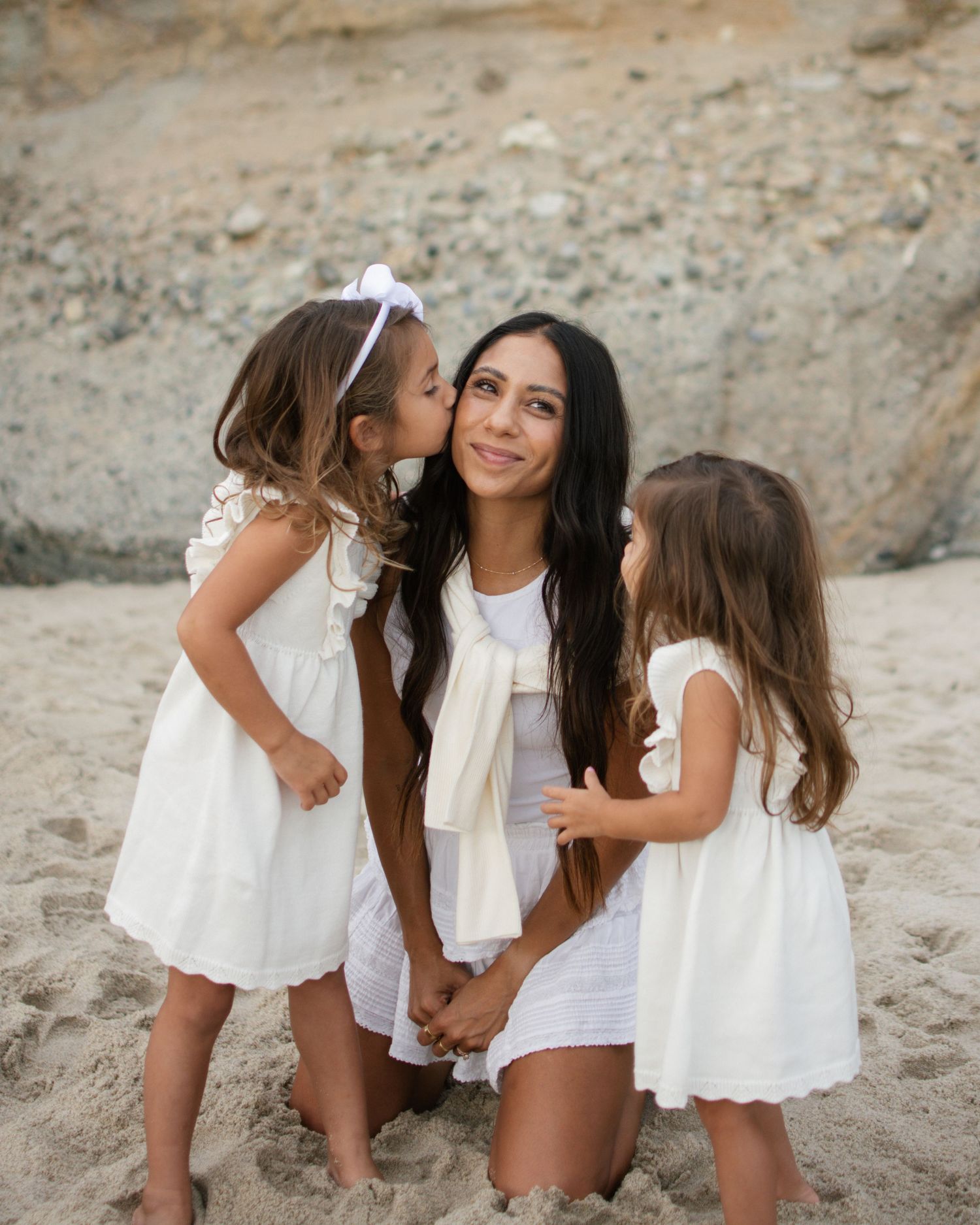 Mother kneeling on beach while two young daughters in white dresses embrace her.