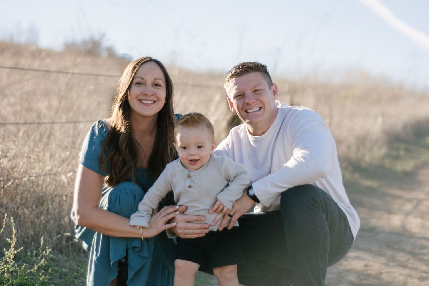 Smiling family of three poses together on a sunlit dirt path surrounded by golden grasses.