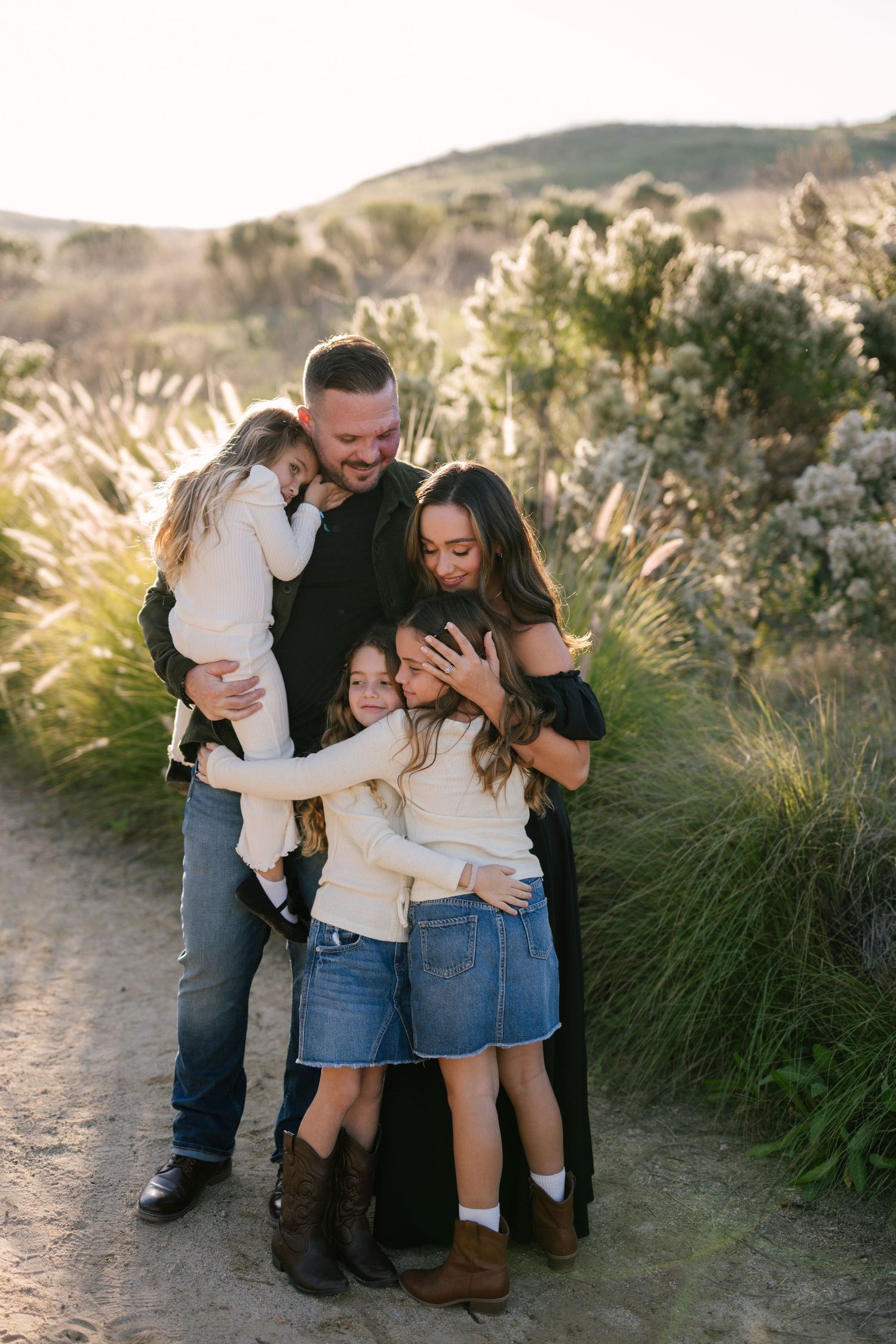 Family of five sharing a warm embrace on a sunlit dirt path surrounded by wild greenery.