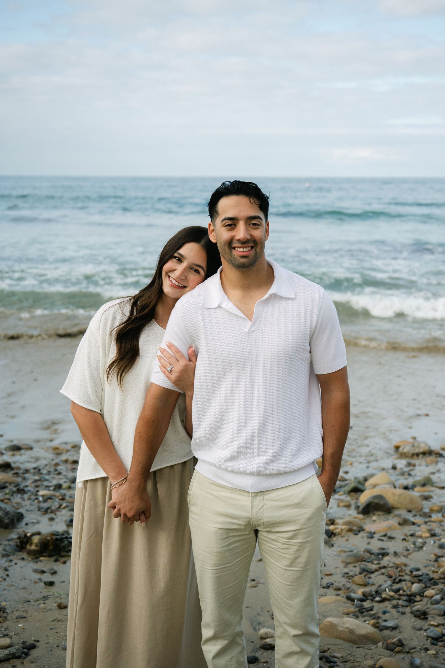 Smiling couple standing together on rocky beach with ocean waves, woman leaning on mans shoulder.