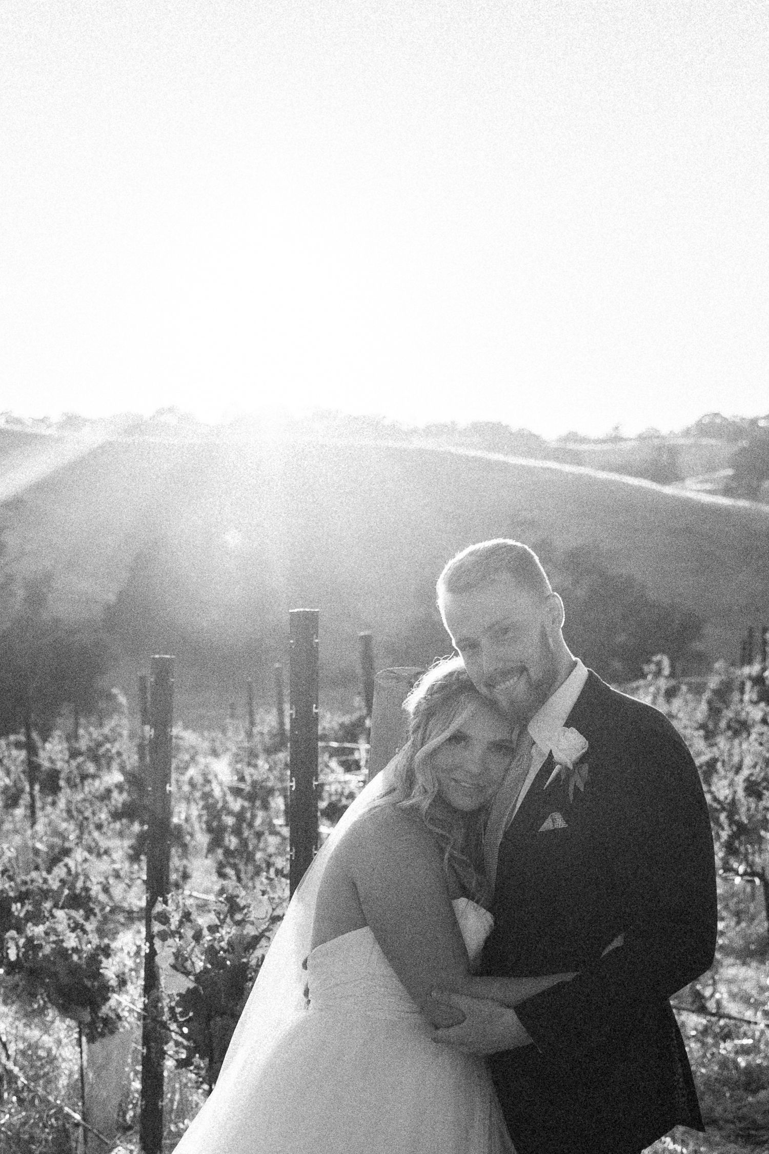 Black and white photo of newlywed couple embracing in sunny vineyard landscape.