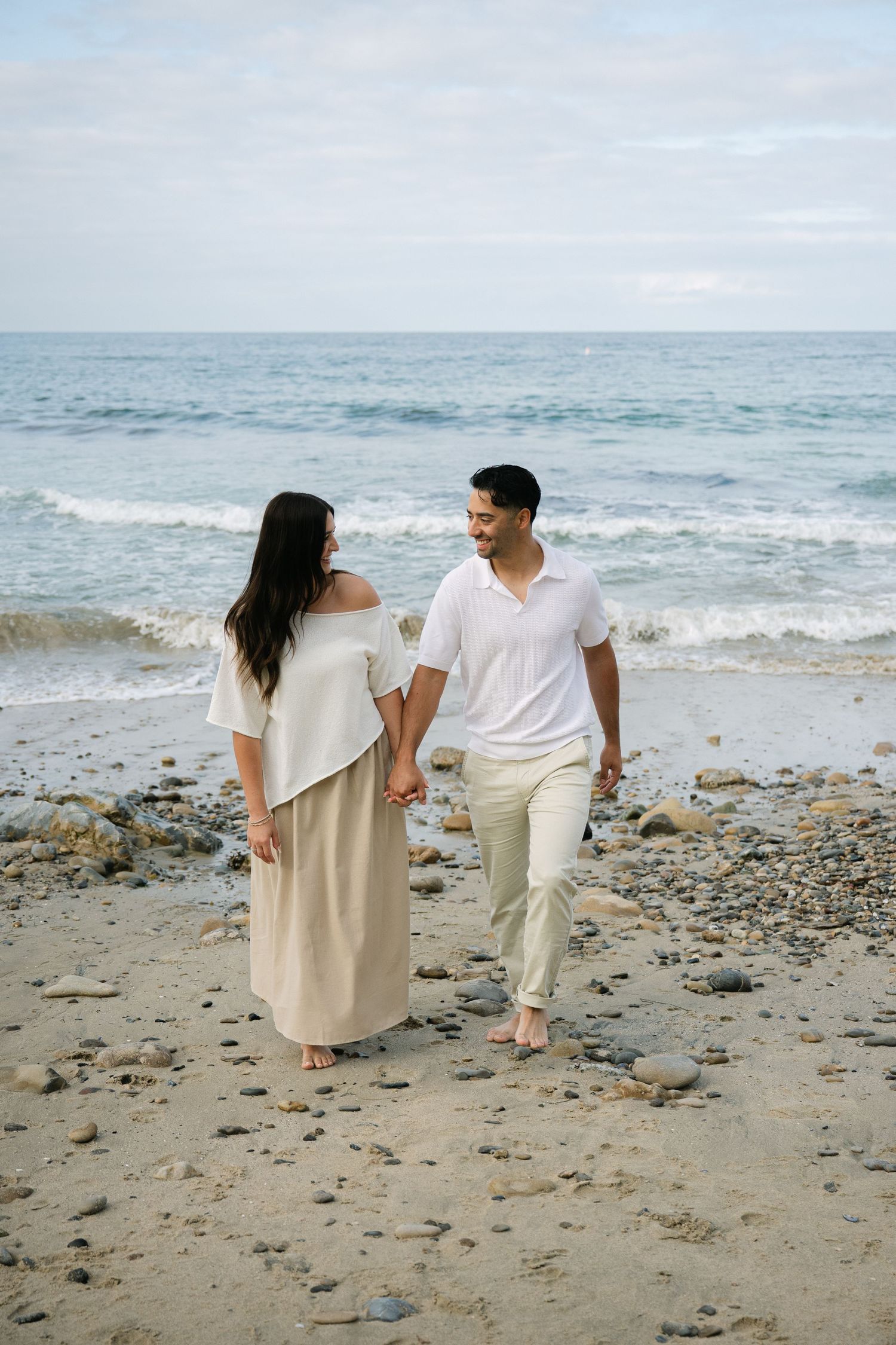 Couple walks hand-in-hand along rocky beach shoreline, smiling at each other with gentle ocean waves in background.