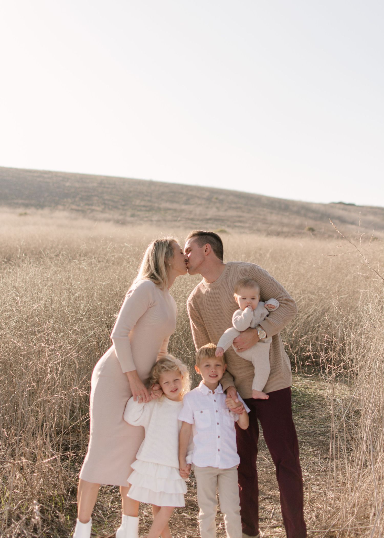 Couple kissing while holding children in a golden field of tall dry grass at sunset.