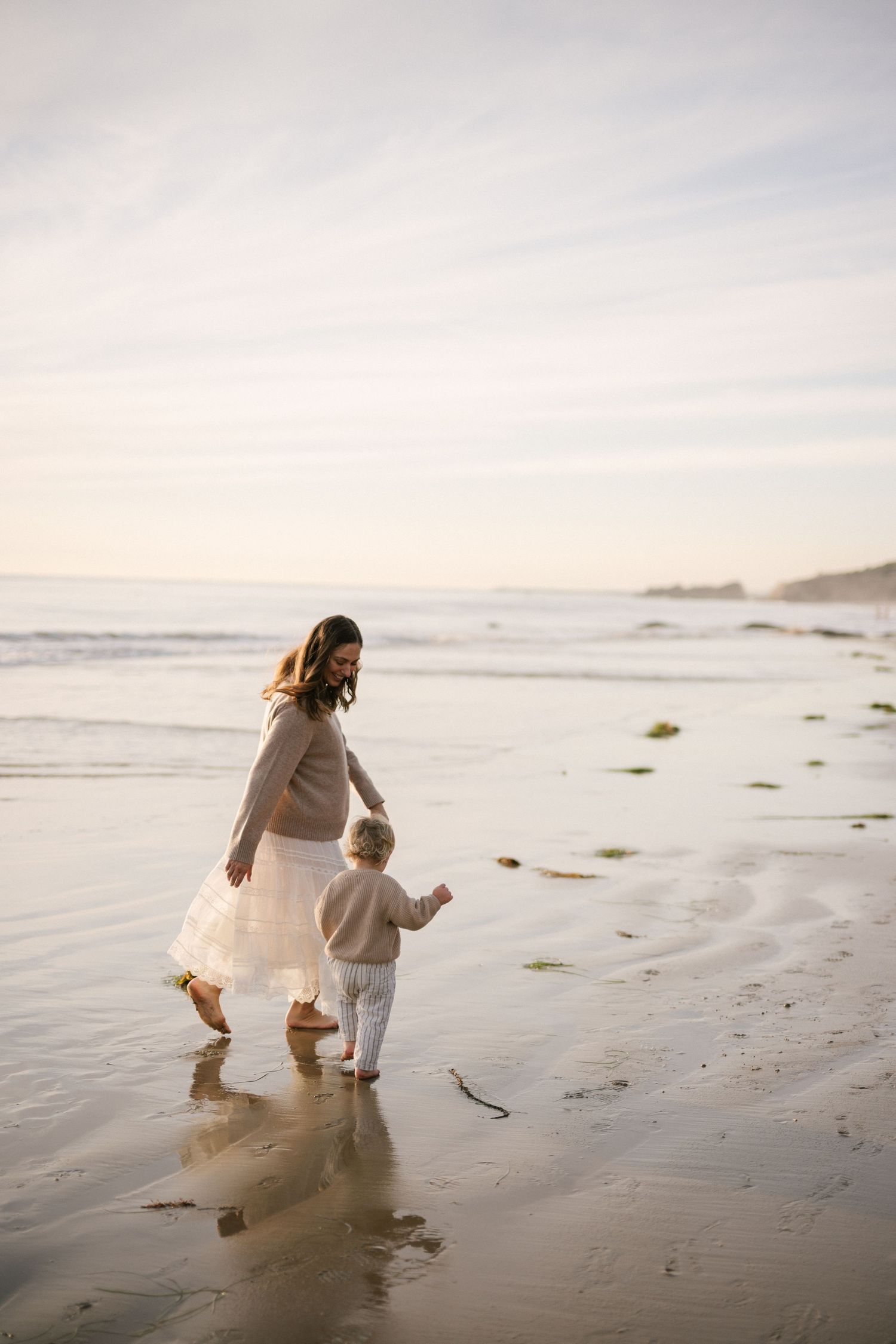Mother in white skirt walking on wet beach sand holding hands with toddler at golden hour.
