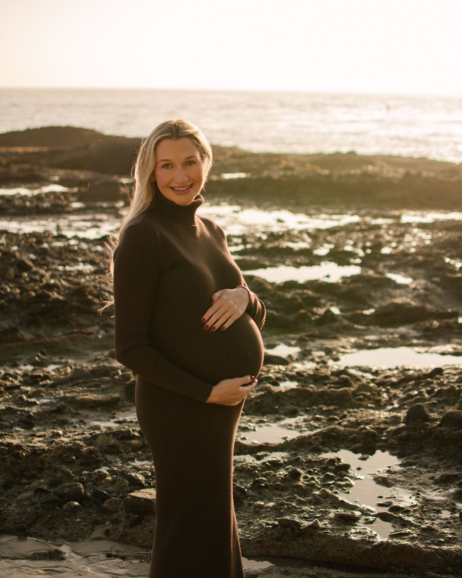 Smiling pregnant woman in dark dress posing at rocky ocean shore.