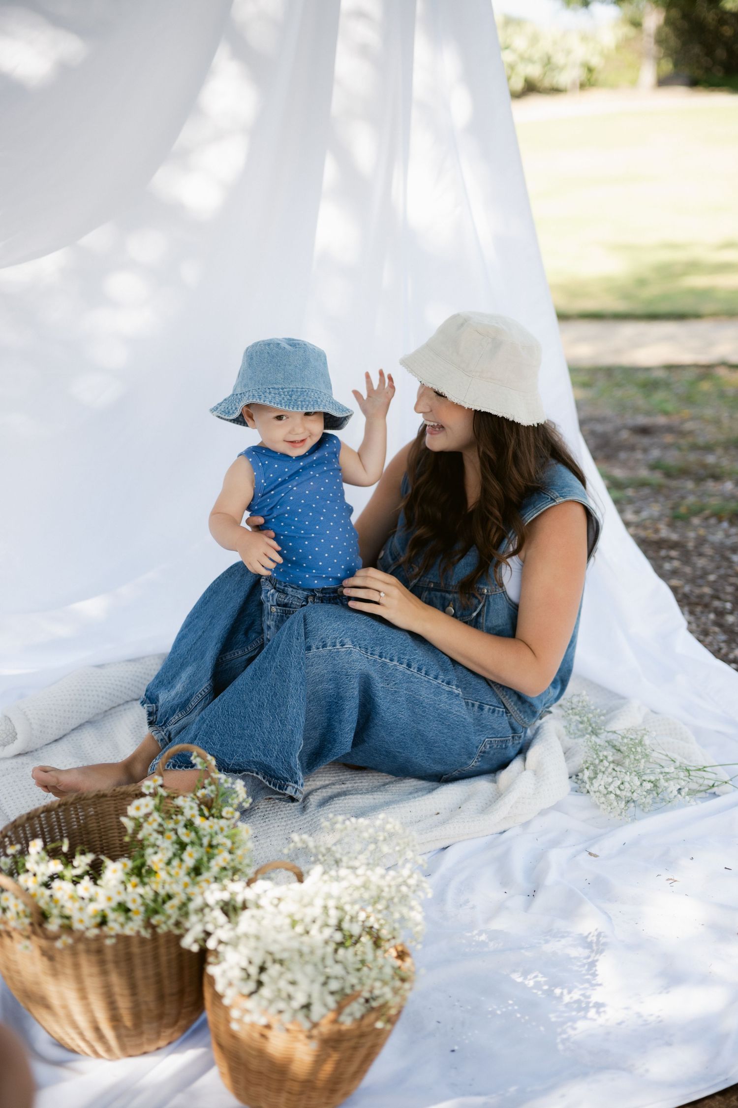 Mother and baby daughter in matching denim outfits and bucket hats at outdoor picnic.