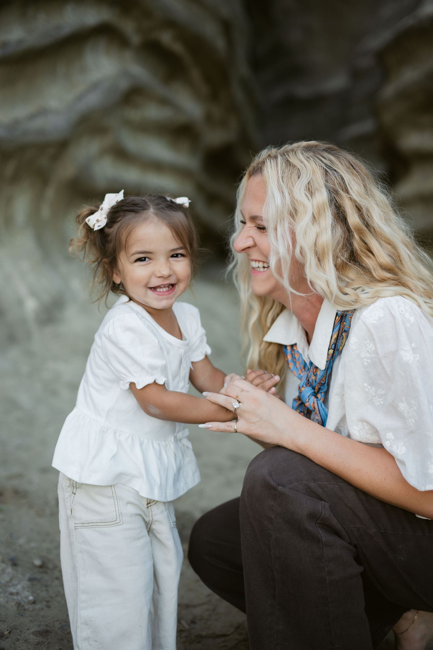 A laughing blonde mother crouches down to interact with her smiling young daughter in a white top outdoors.