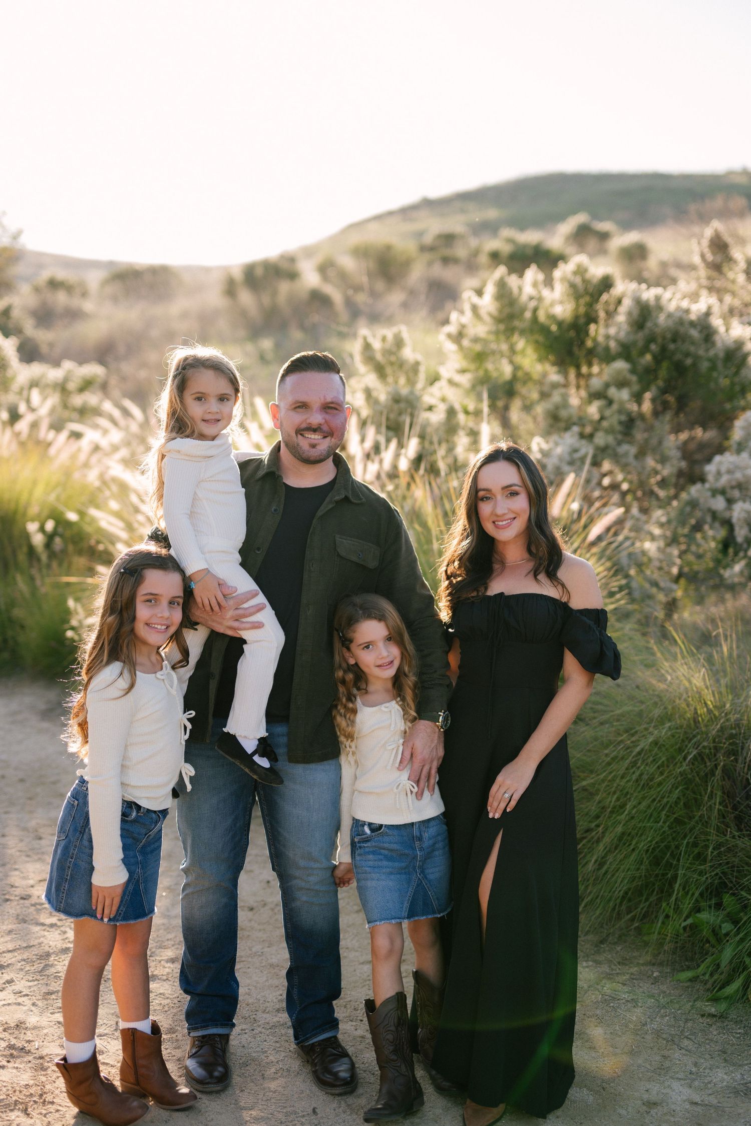 Family of five posing together on a sunny trail with lush green shrubs in the background.