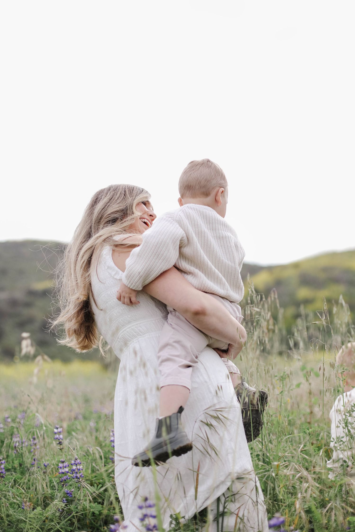 Mother in white dress twirling with her baby in a lush green field of wildflowers.