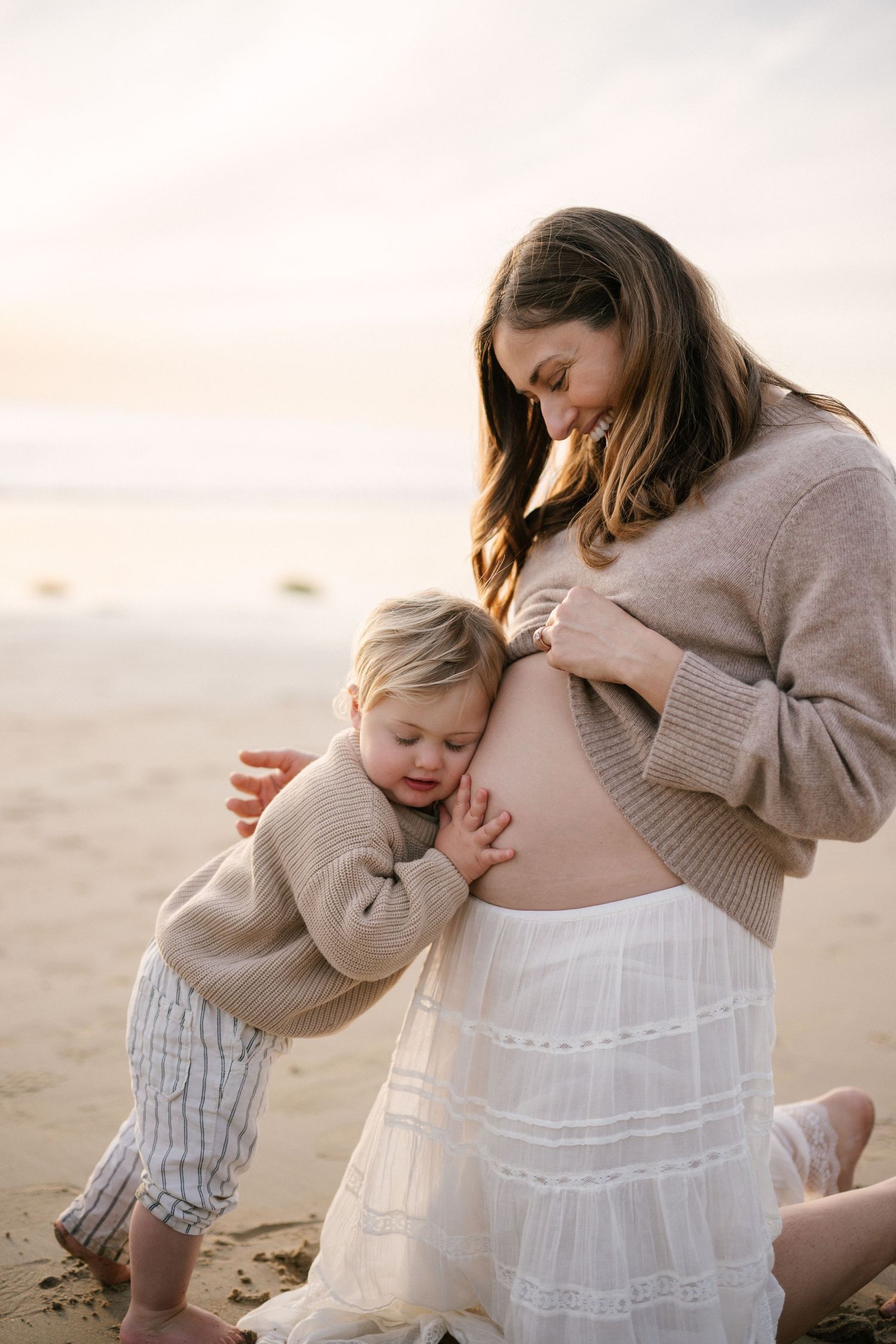 Pregnant woman at beach sunset with toddler hugging her baby bump.