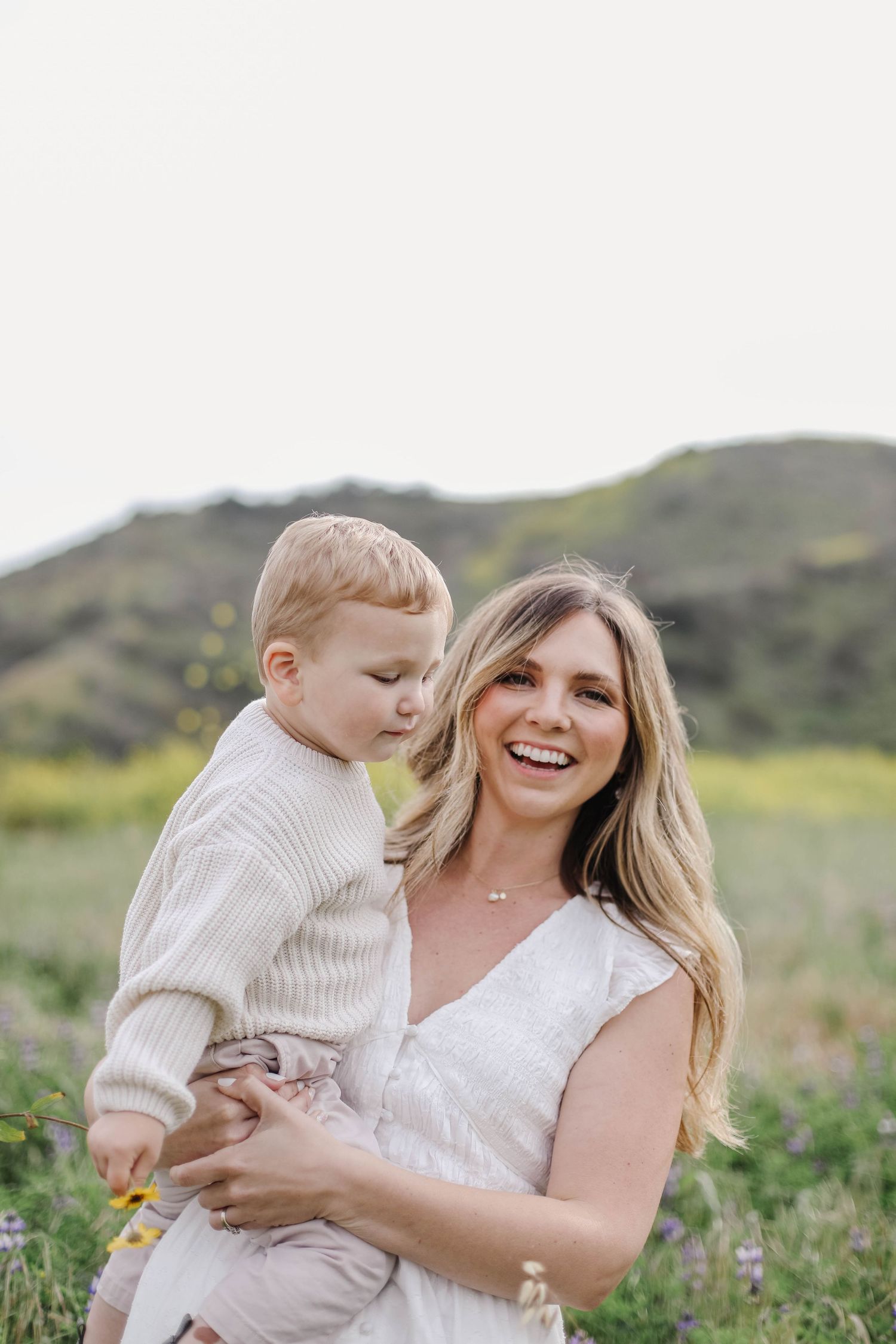 Smiling mother holding her toddler son in a sunny wildflower meadow with green hills in the background.
