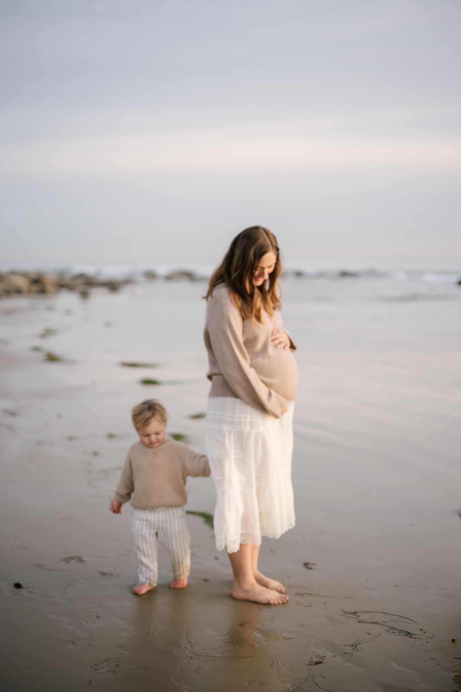Pregnant woman in beige sweater holding toddlers hand on sandy beach.
