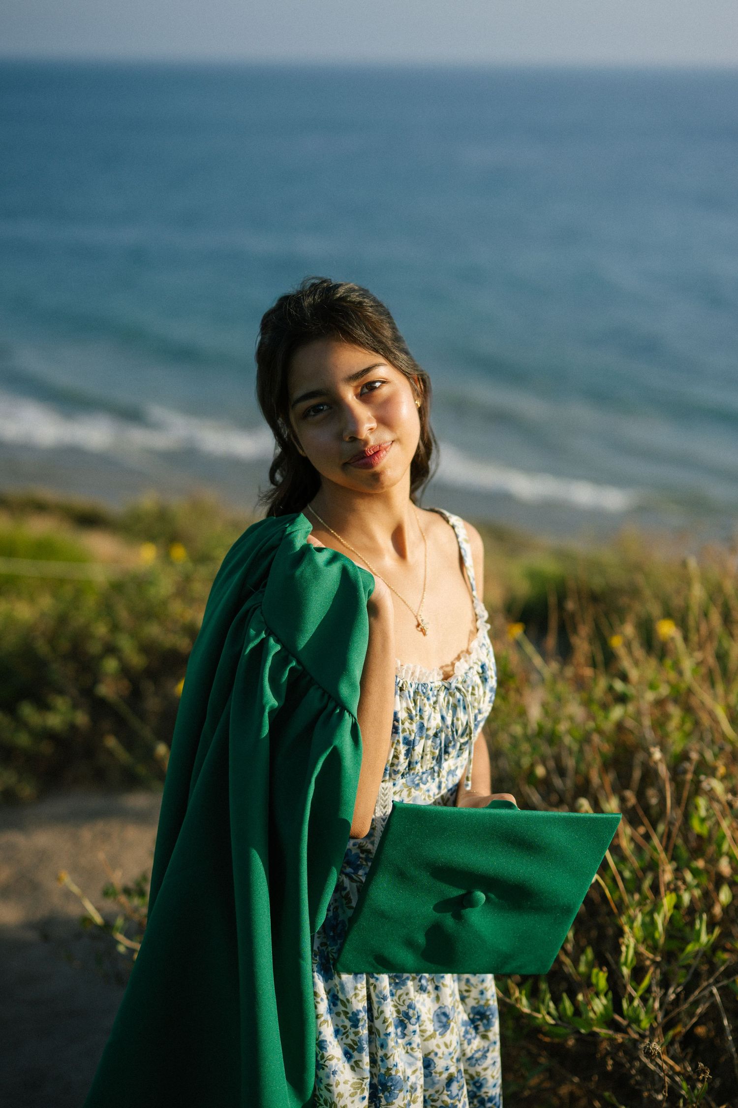 Young woman in green graduation gown posing near the ocean at sunset.