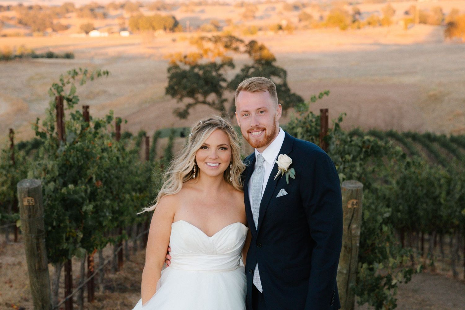 Happy newlyweds standing together in front of lush vineyard rows at sunset.