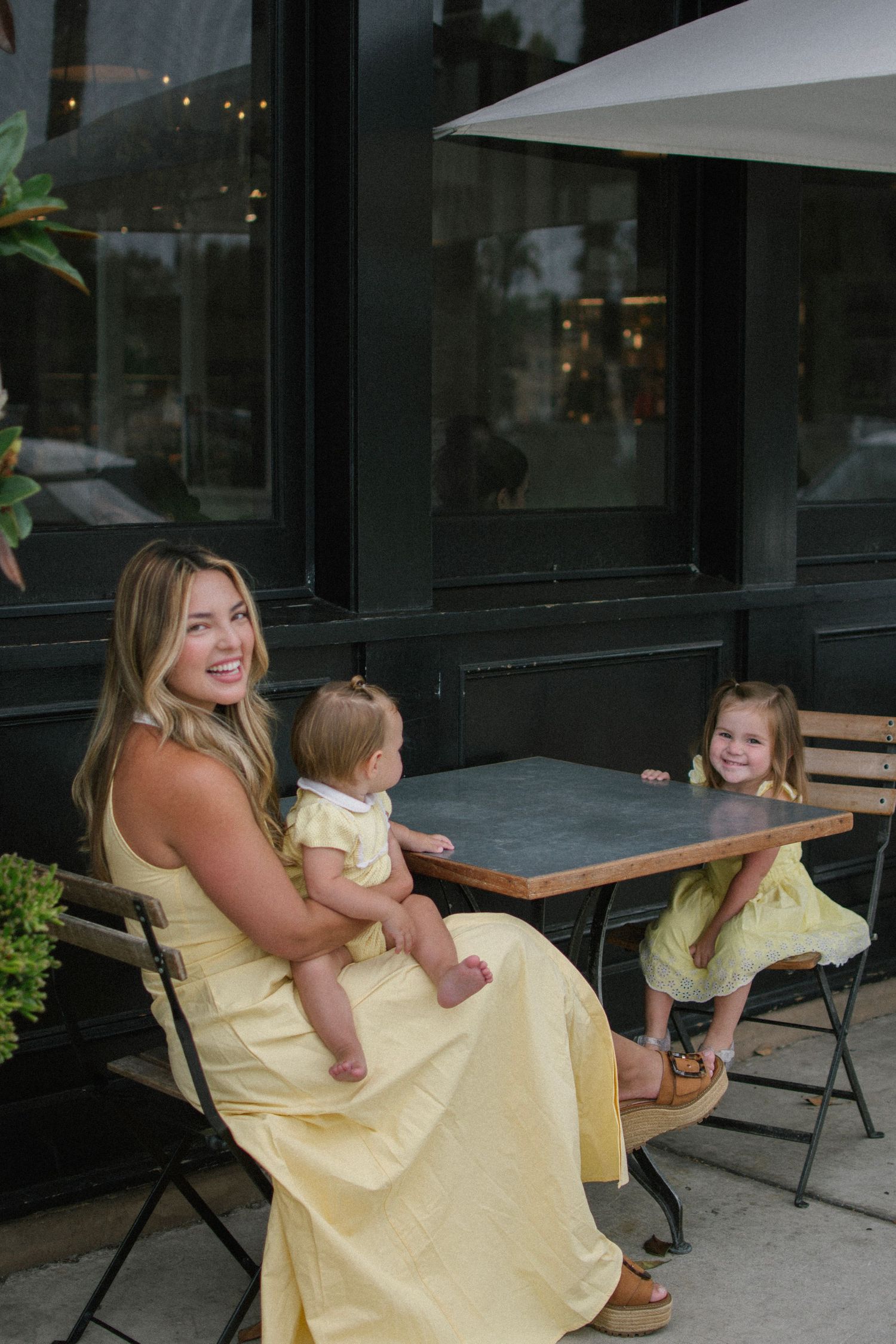 A blonde mother in a yellow dress sits at an outdoor cafe table with her baby and young daughter.