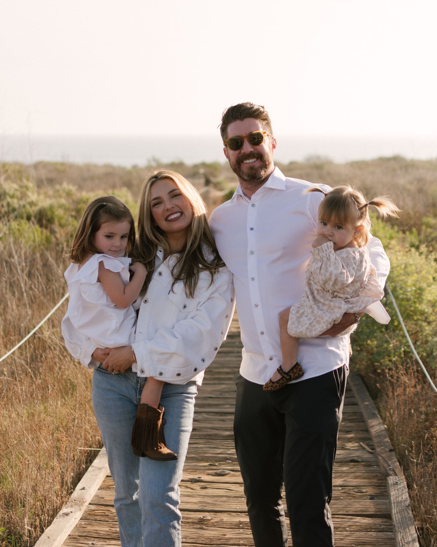 Smiling family of four posing on wooden boardwalk, parents each holding a young daughter outdoors.