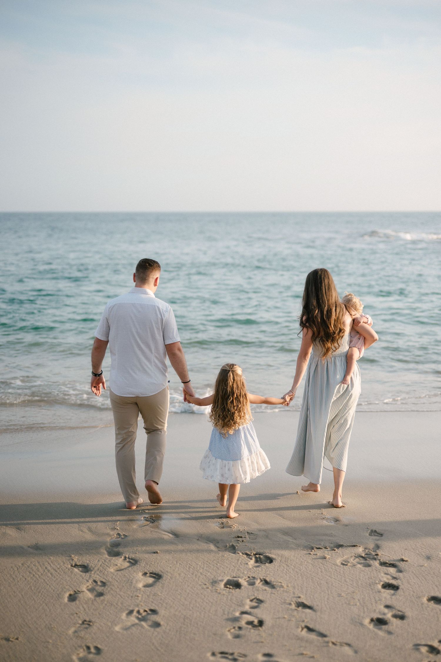 Family of four walking hand-in-hand toward turquoise ocean on sandy beach leaving footprints behind.