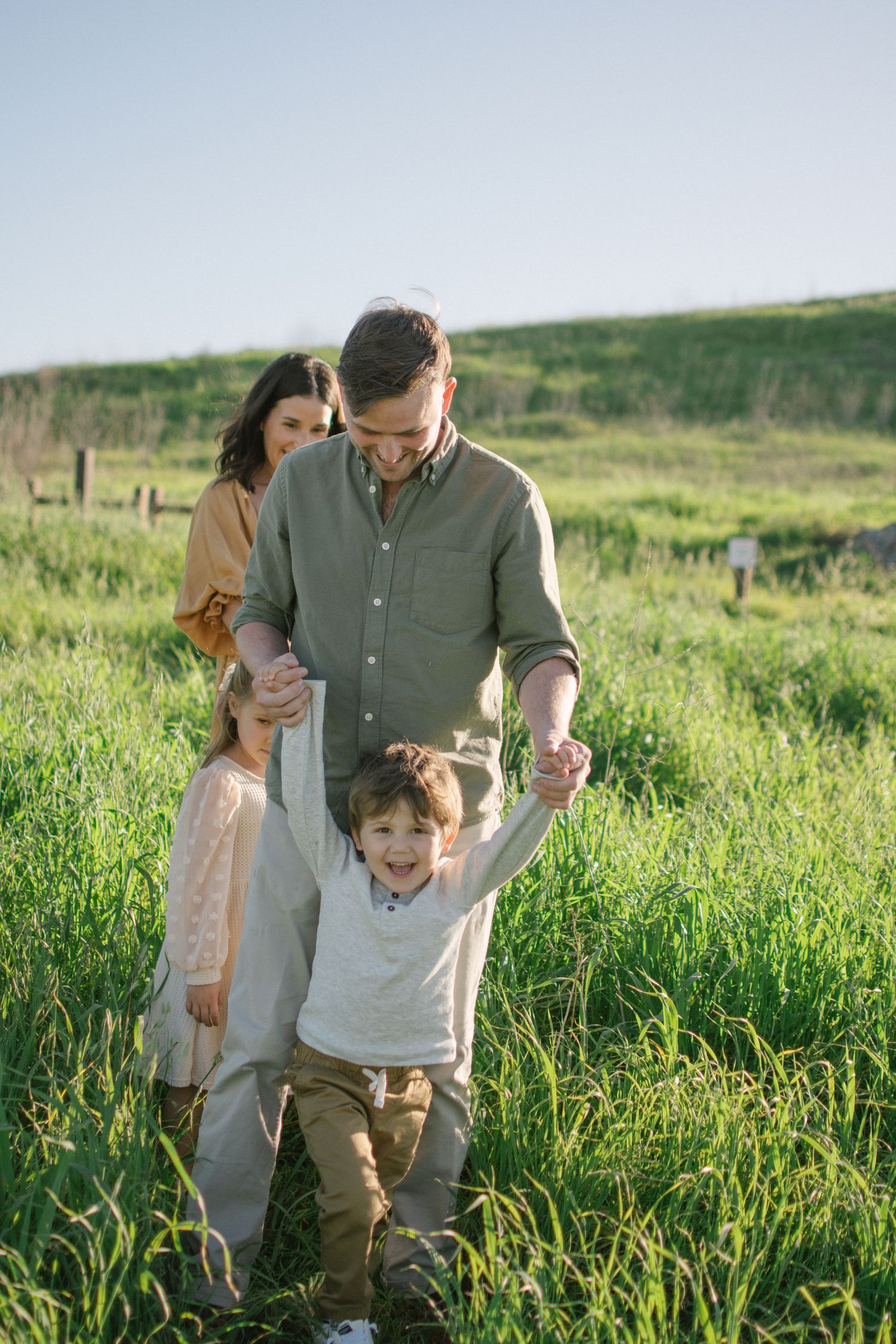 Smiling father swings joyful boy by the hands while mother watches in a green meadow.