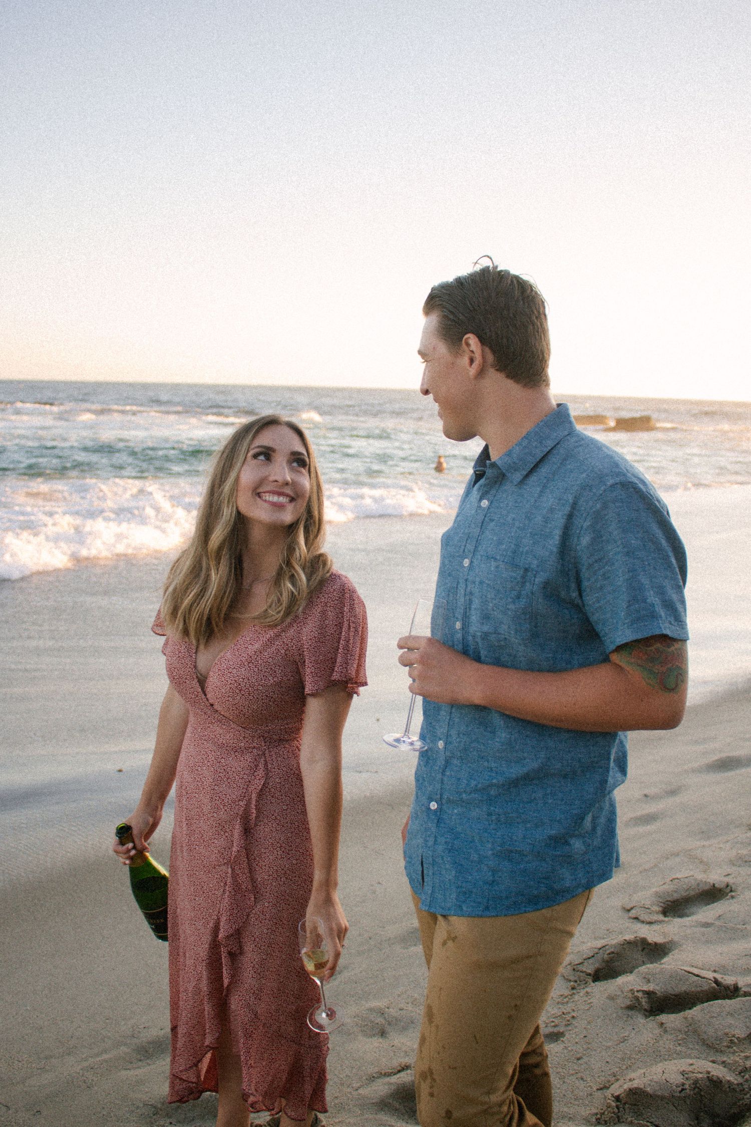 Couple strolling on a beach at golden hour, laughing and holding champagne glasses near the shoreline.