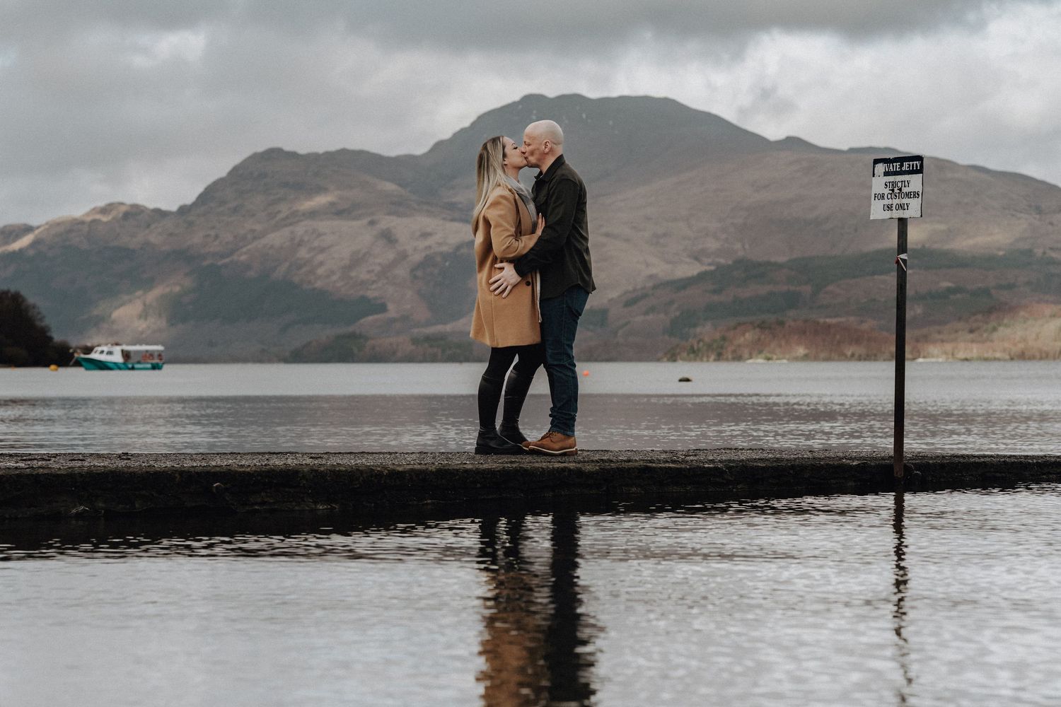 Couple embracing on a pier with Scottish highlands and moody skies in the background.