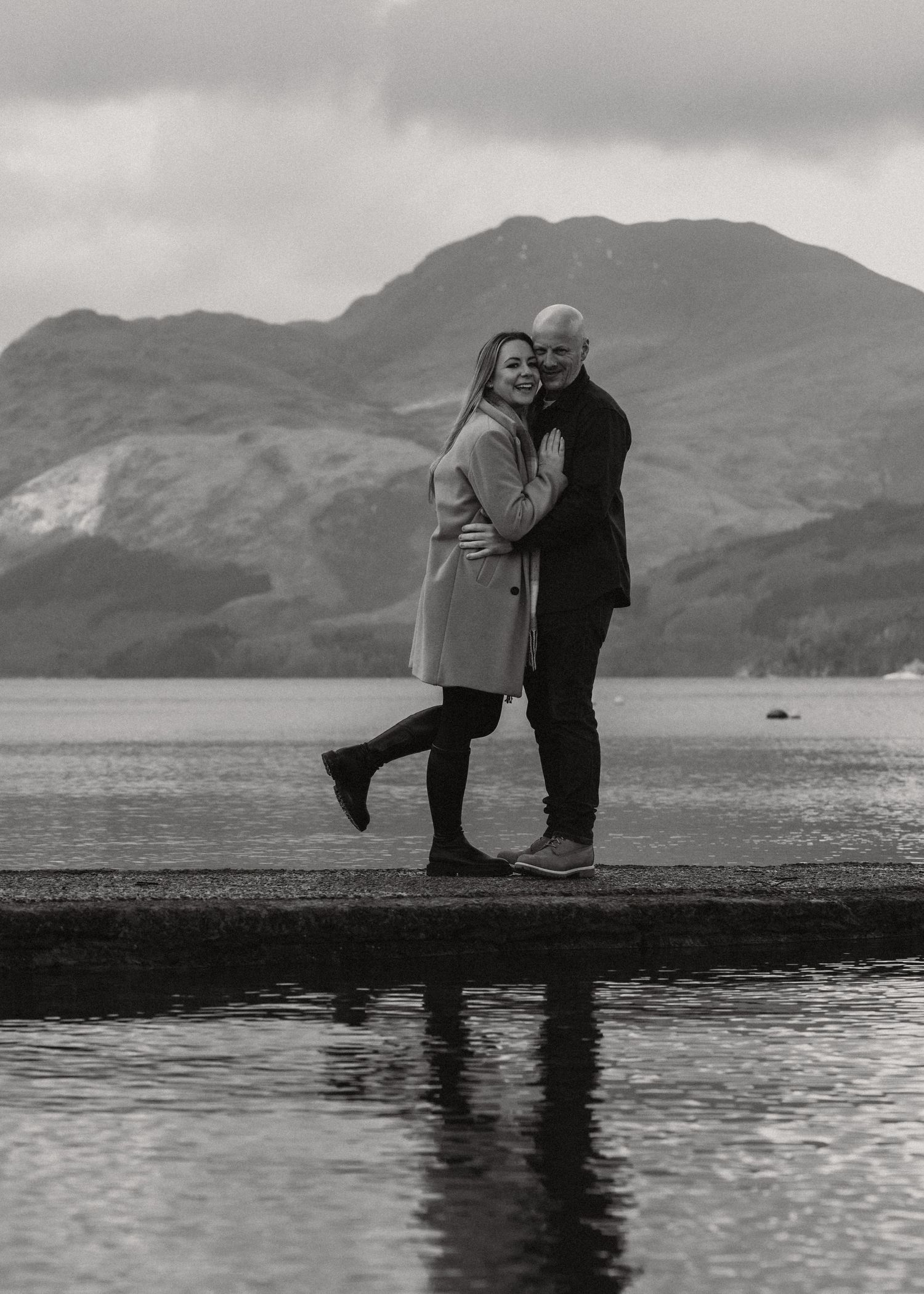 Black and white photo of a happy couple on a lakeside pier with mountains in the distance.