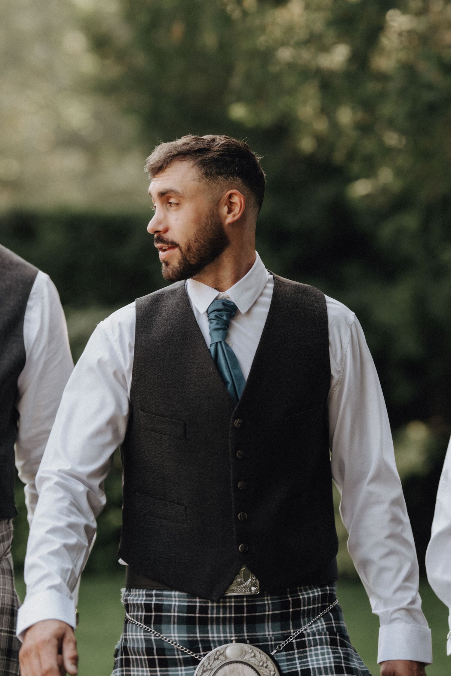 Groomsman in traditional Scottish wedding attire with charcoal vest, white shirt and plaid kilt at outdoor celebration.