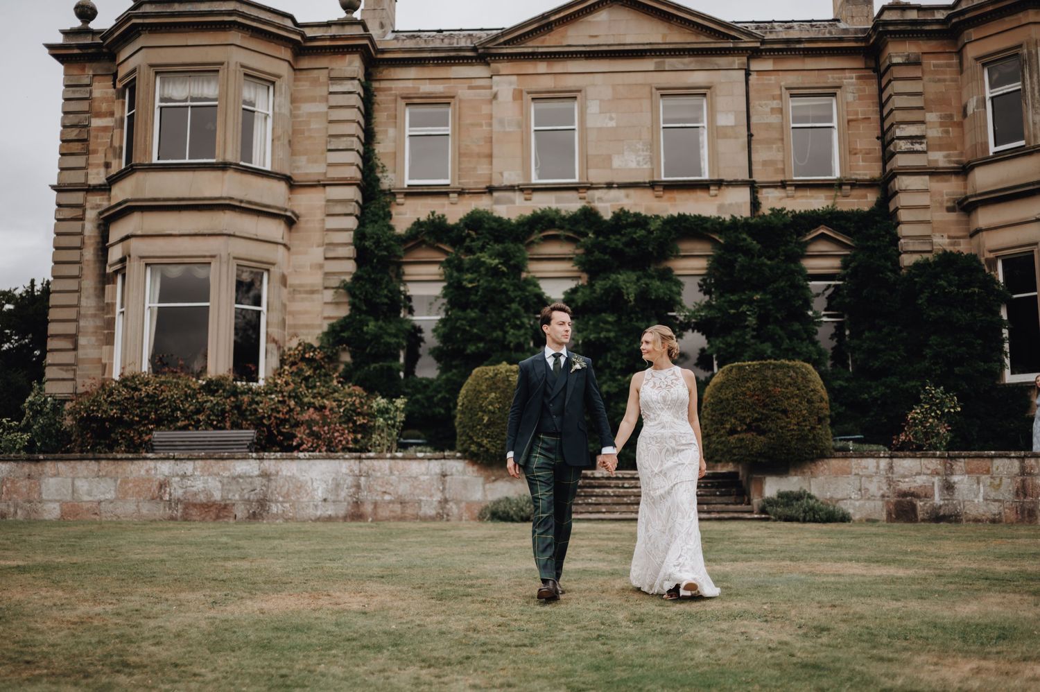 A wedding couple walking outdoors near a historic stone manor with grand architecture visible in the background.