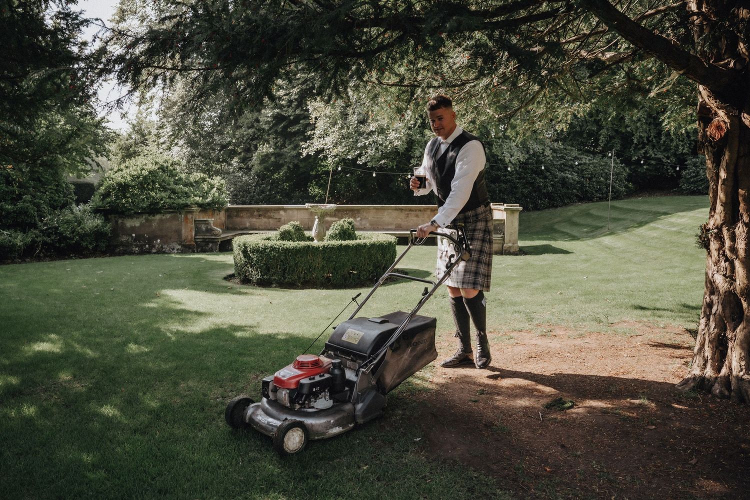 A person in traditional Scottish attire mows a lawn in a manicured garden setting.