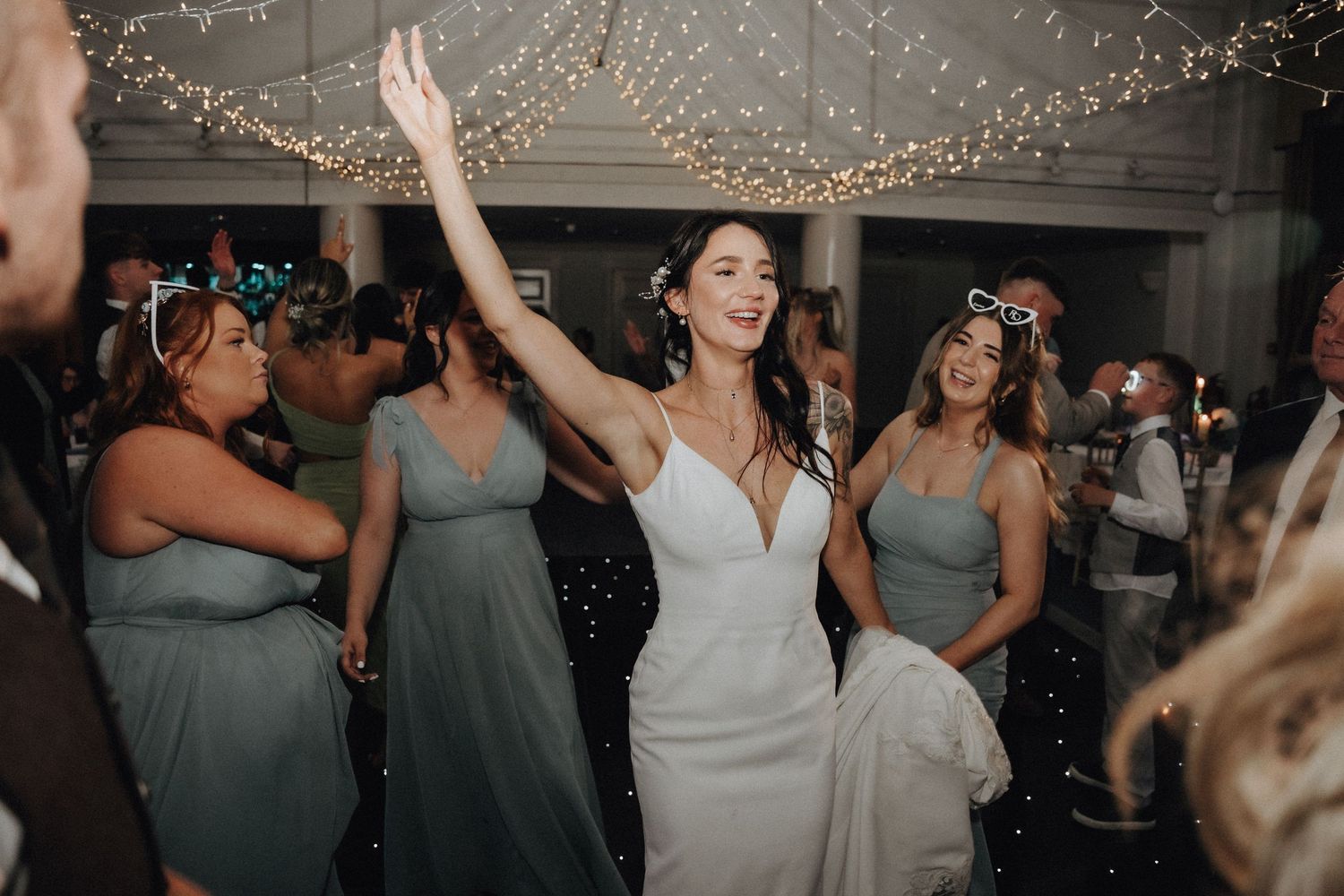 Bride dancing joyfully at reception with bridesmaids in sage green dresses.