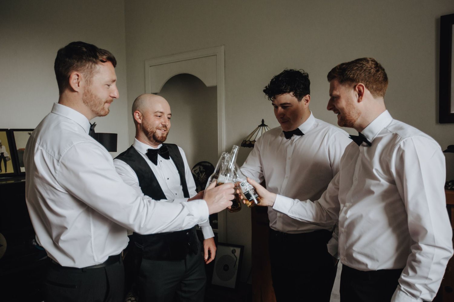 Men in white dress shirts gather to help pin a buttonhole flower to a formal outfit in a candid moment.