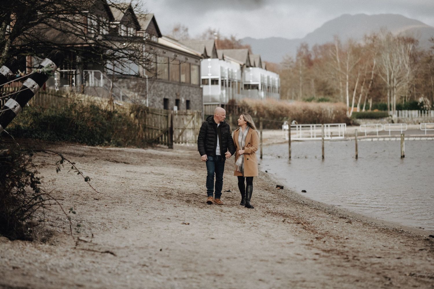 A couple walks along a lakeside path with Victorian houses visible in the background.