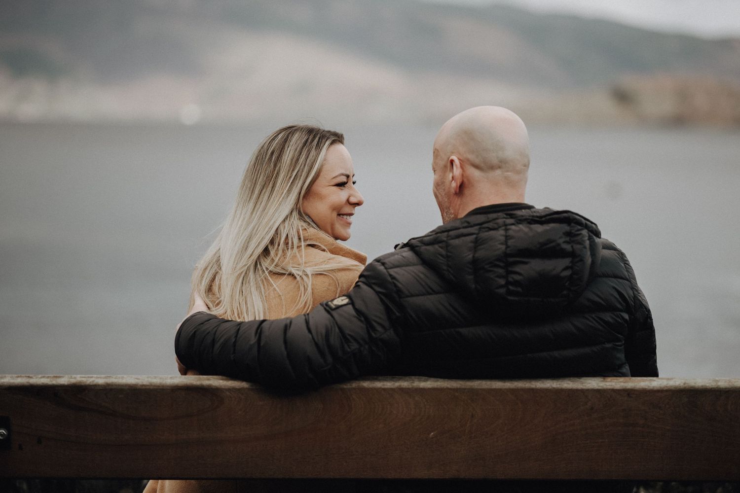 A couple sharing a tender moment on a wooden bench overlooking a misty lake and mountains.
