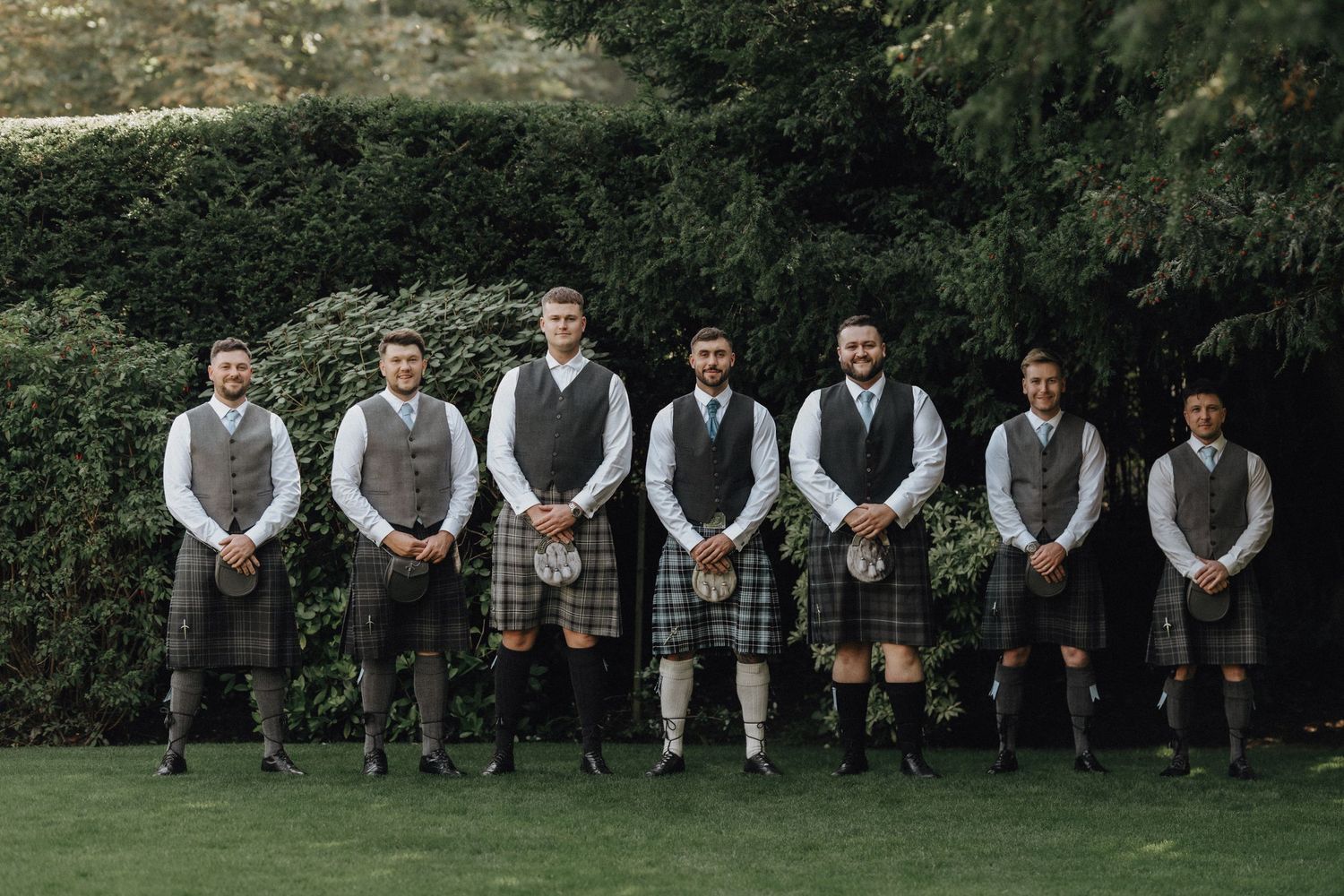 Groomsmen in matching waistcoats and kilts stand in line for a formal outdoor portrait.