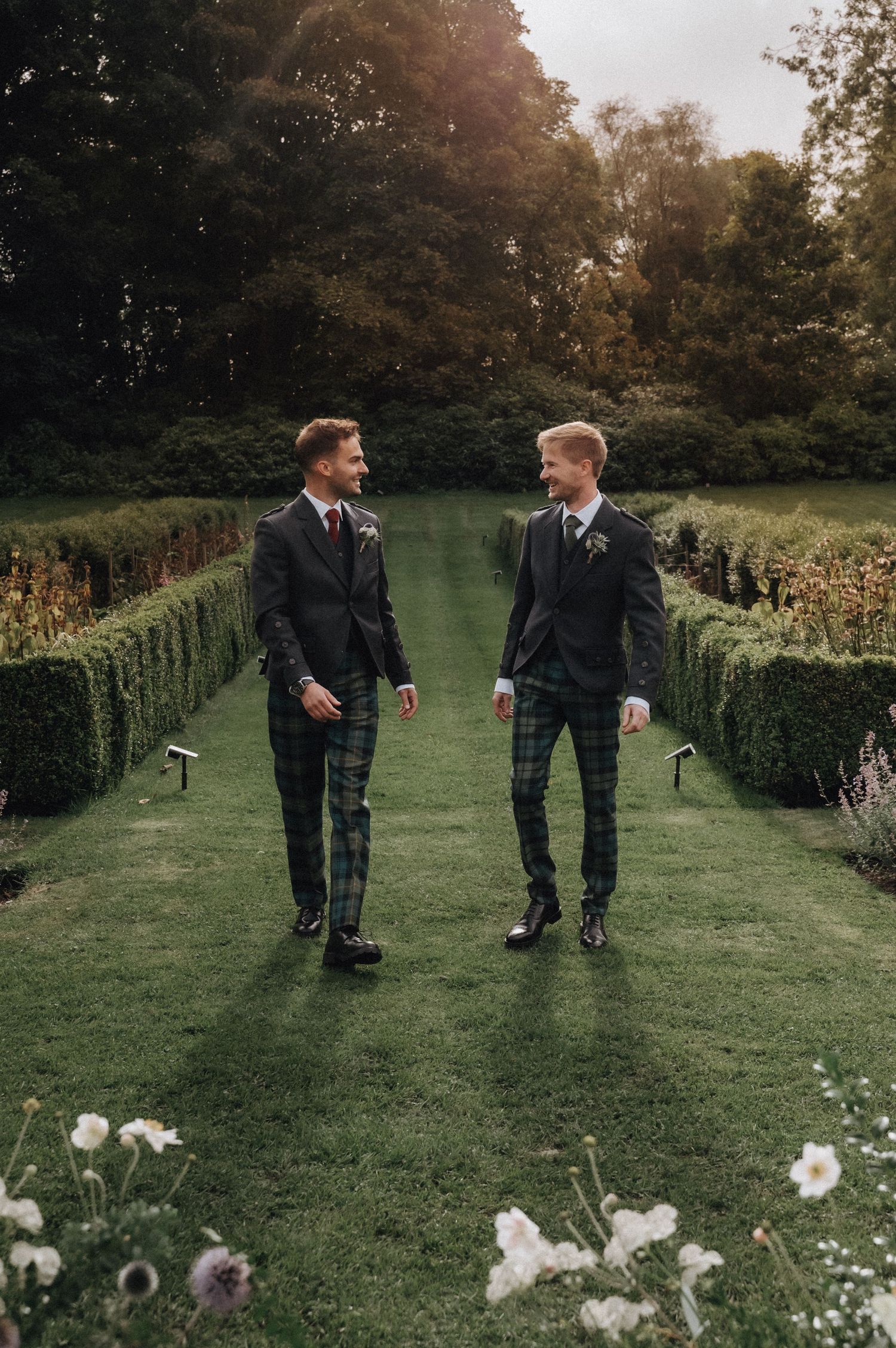 Two people in matching suits and kilts walking through a formal garden with hedges.