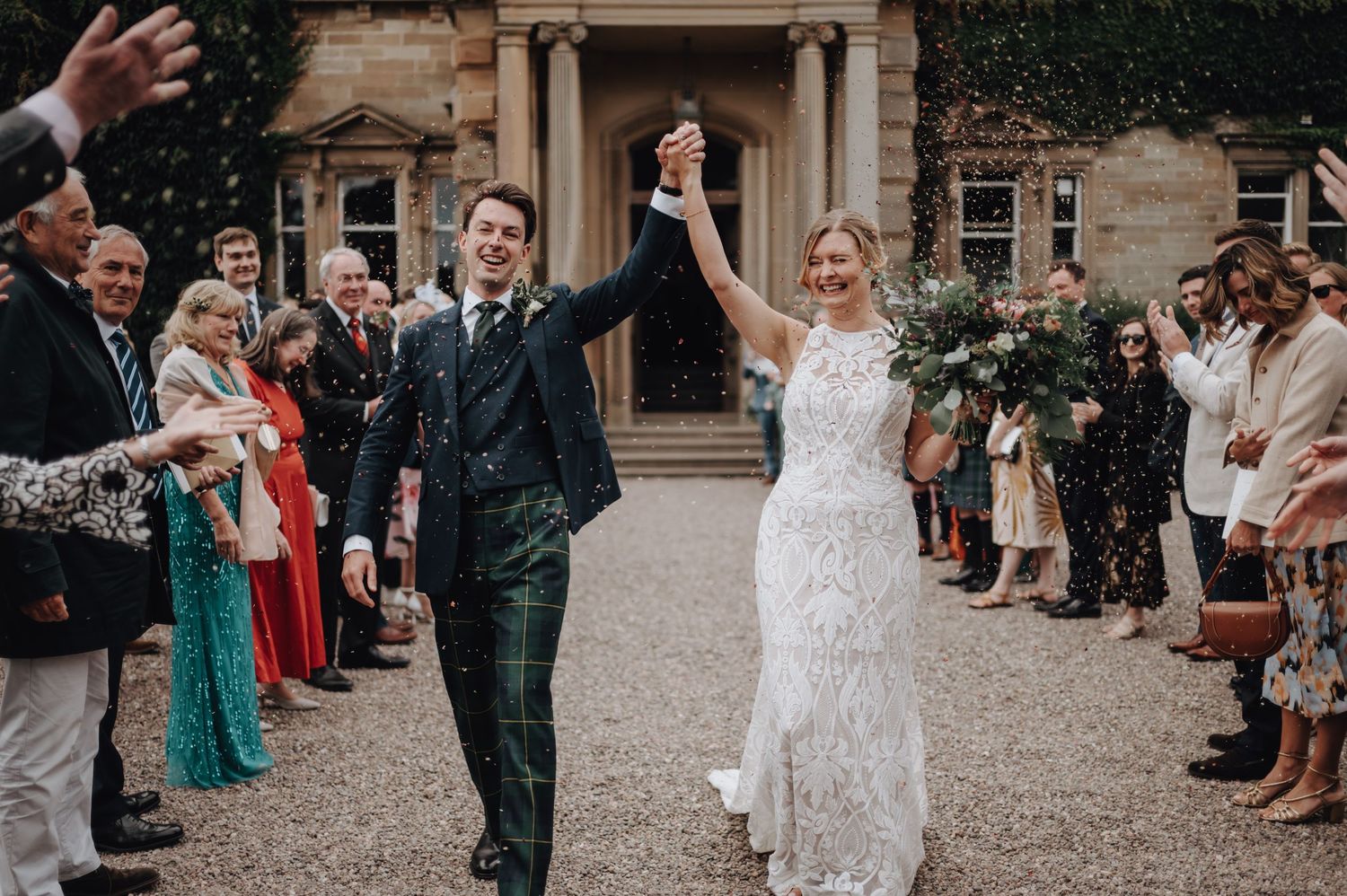A happy couple celebrates with wedding guests by holding hands and raising arms victoriously during their outdoor celebration.