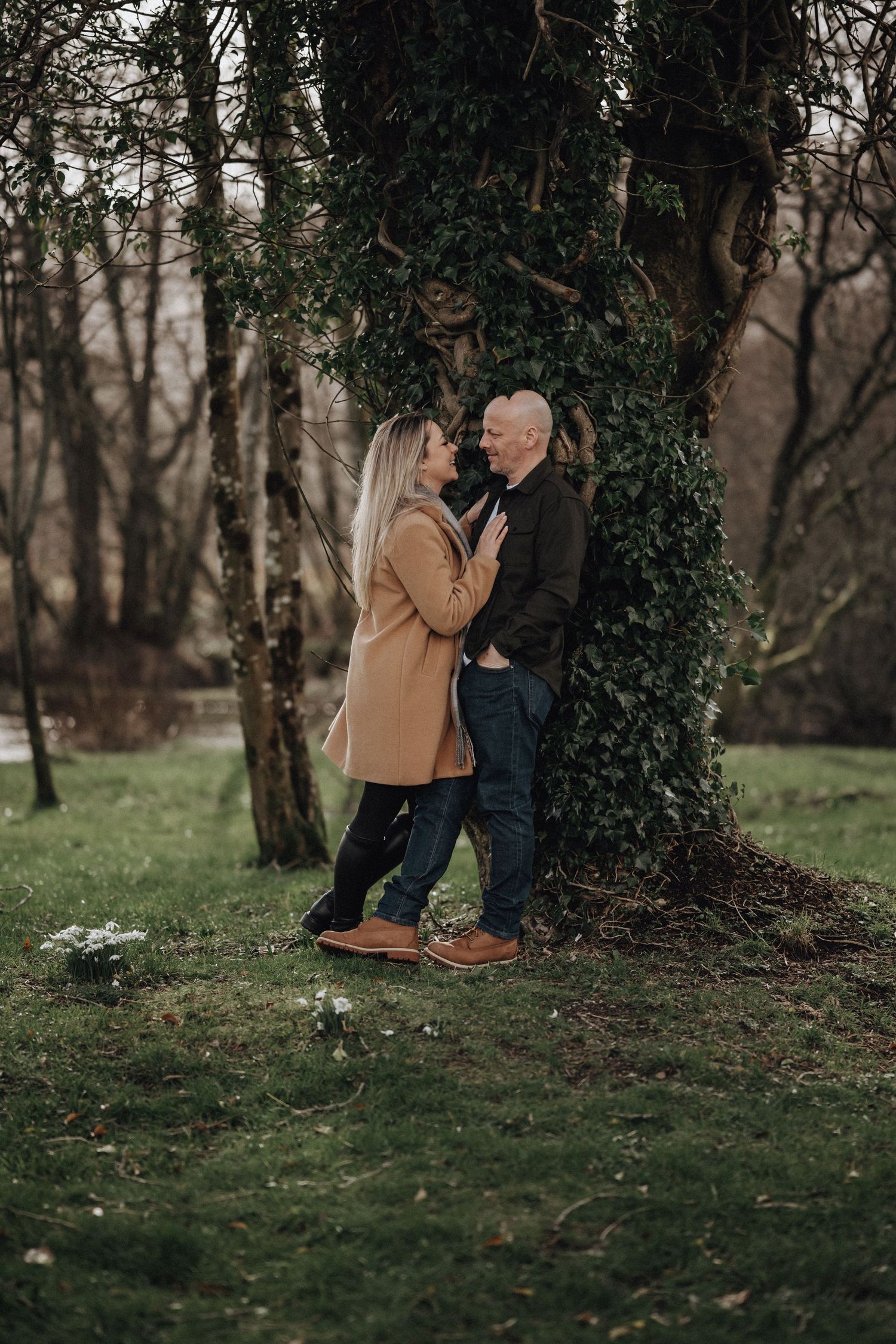 Couple sharing romantic moment against ivy-covered tree in woodland setting.