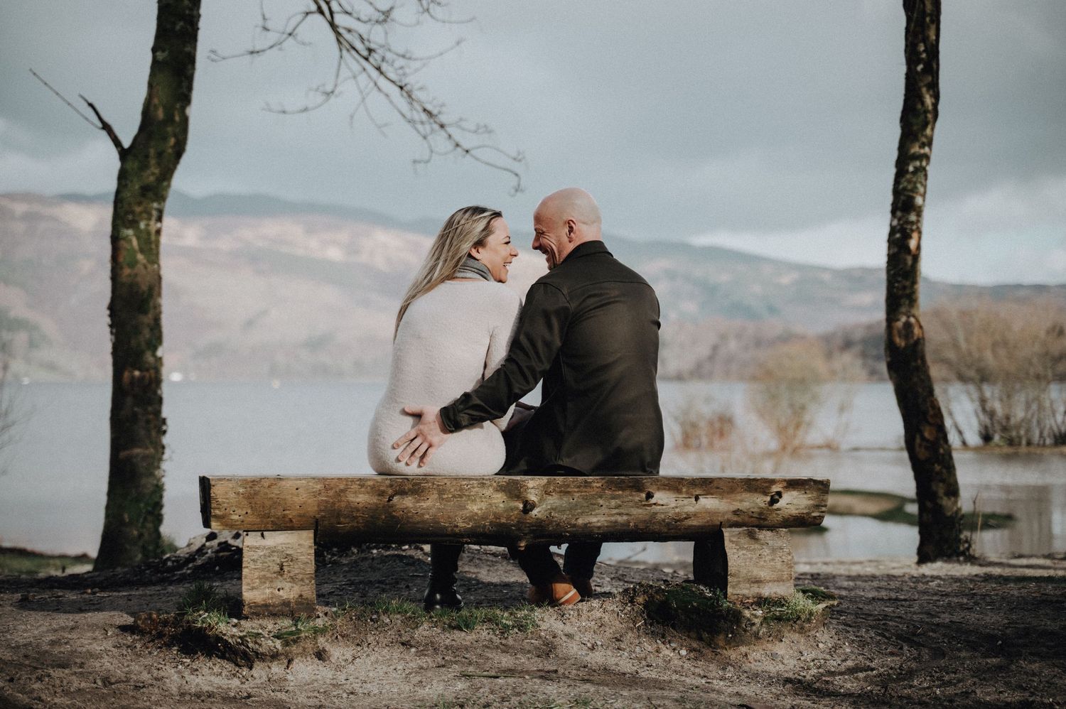 Couple sits together on rustic wooden bench overlooking misty lake and mountains.