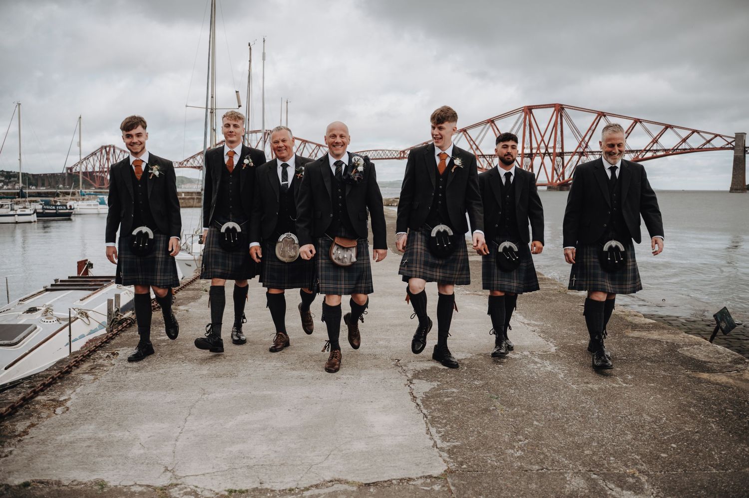 Groomsmen in traditional Scottish kilts walking along harbor with Forth Bridge in background.