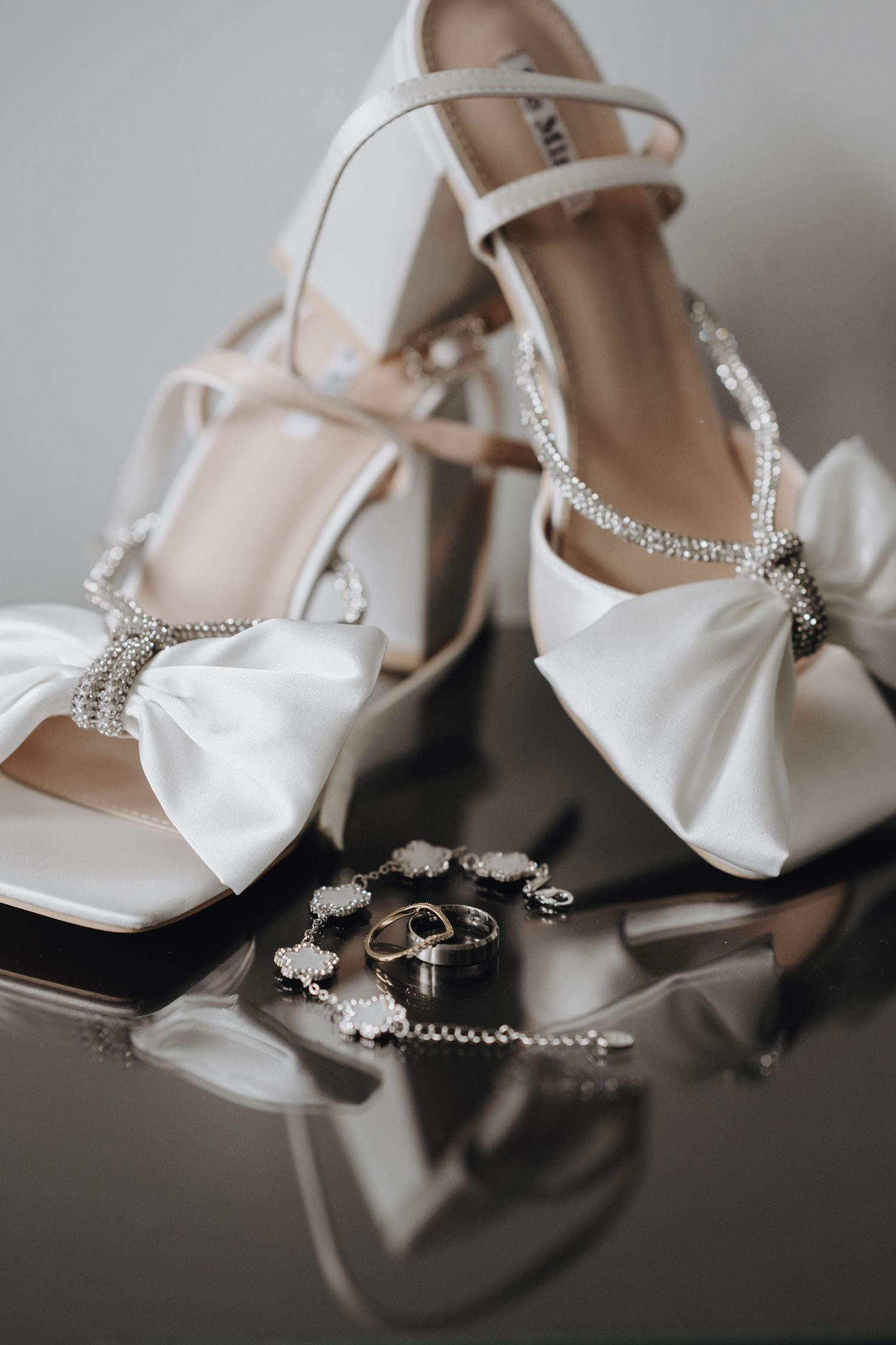 Wedding accessories including white satin bridal shoes with crystal bows and delicate jewelry arranged on reflective surface.