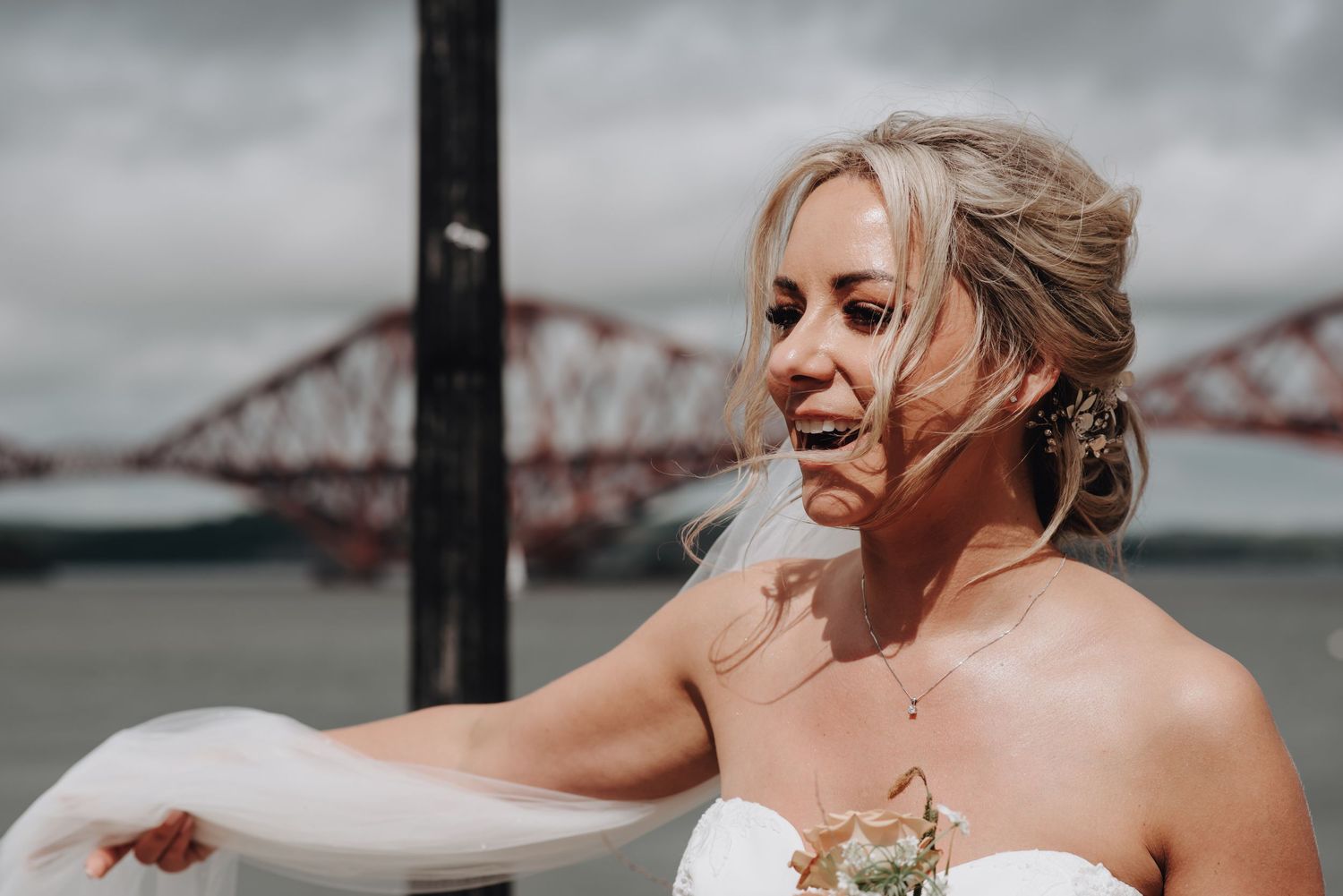 Bride in strapless white gown smiles with Forth Bridge visible in background on overcast day.