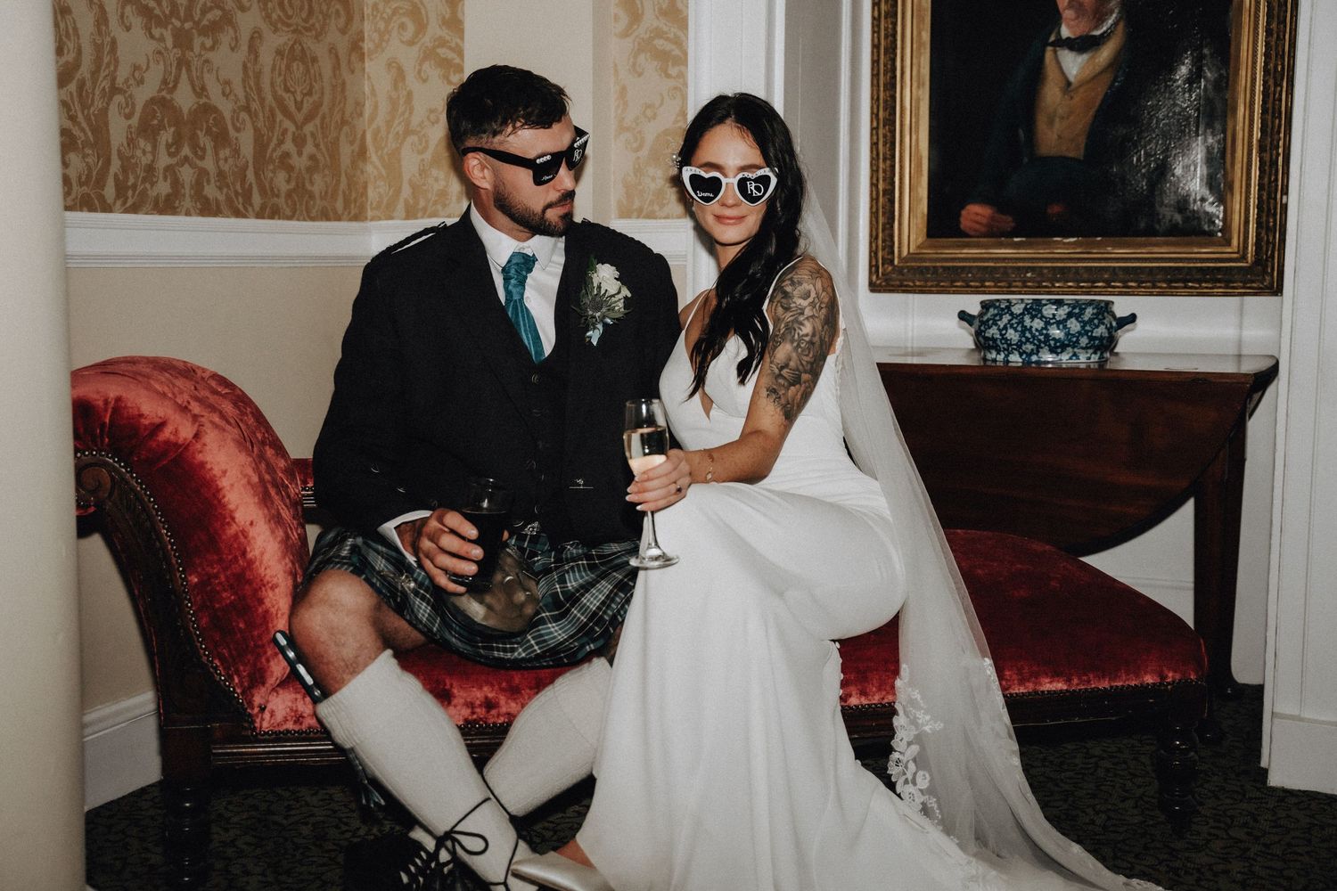 Bride and groom in traditional Scottish wedding attire pose together on vintage red couch.