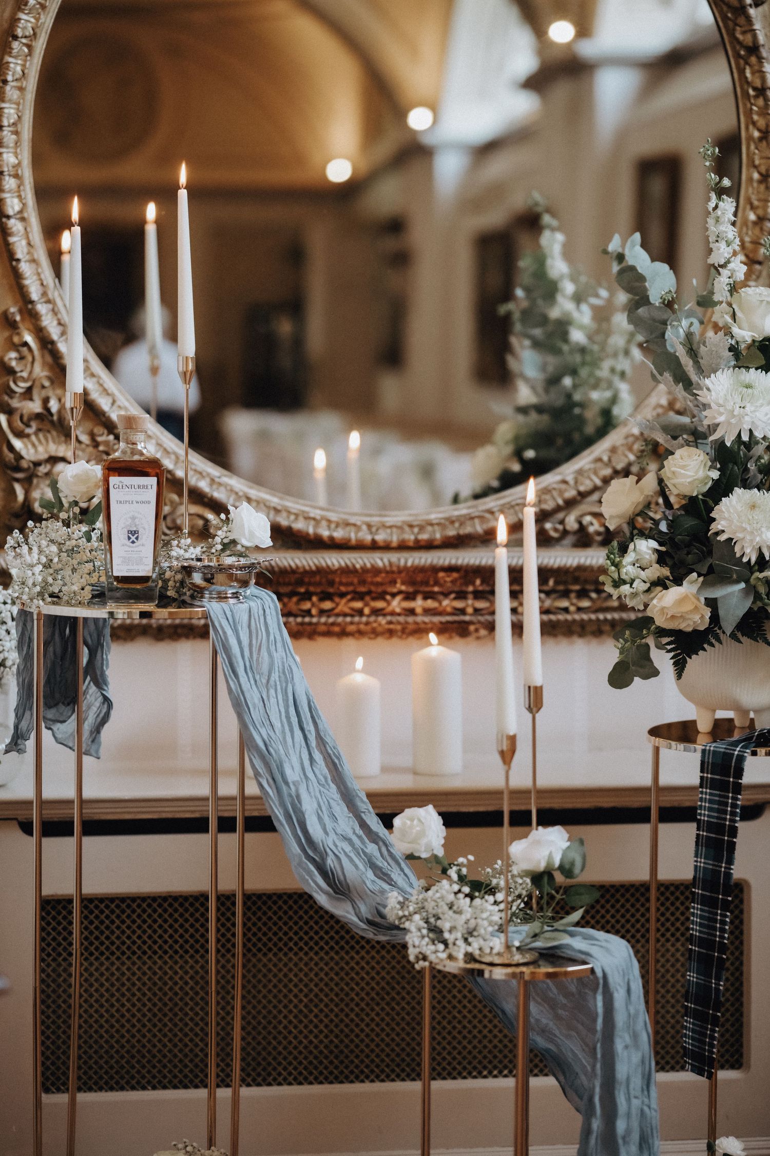 Elegant wedding decor with candles, blue fabric and floral arrangements reflected in ornate mirror.