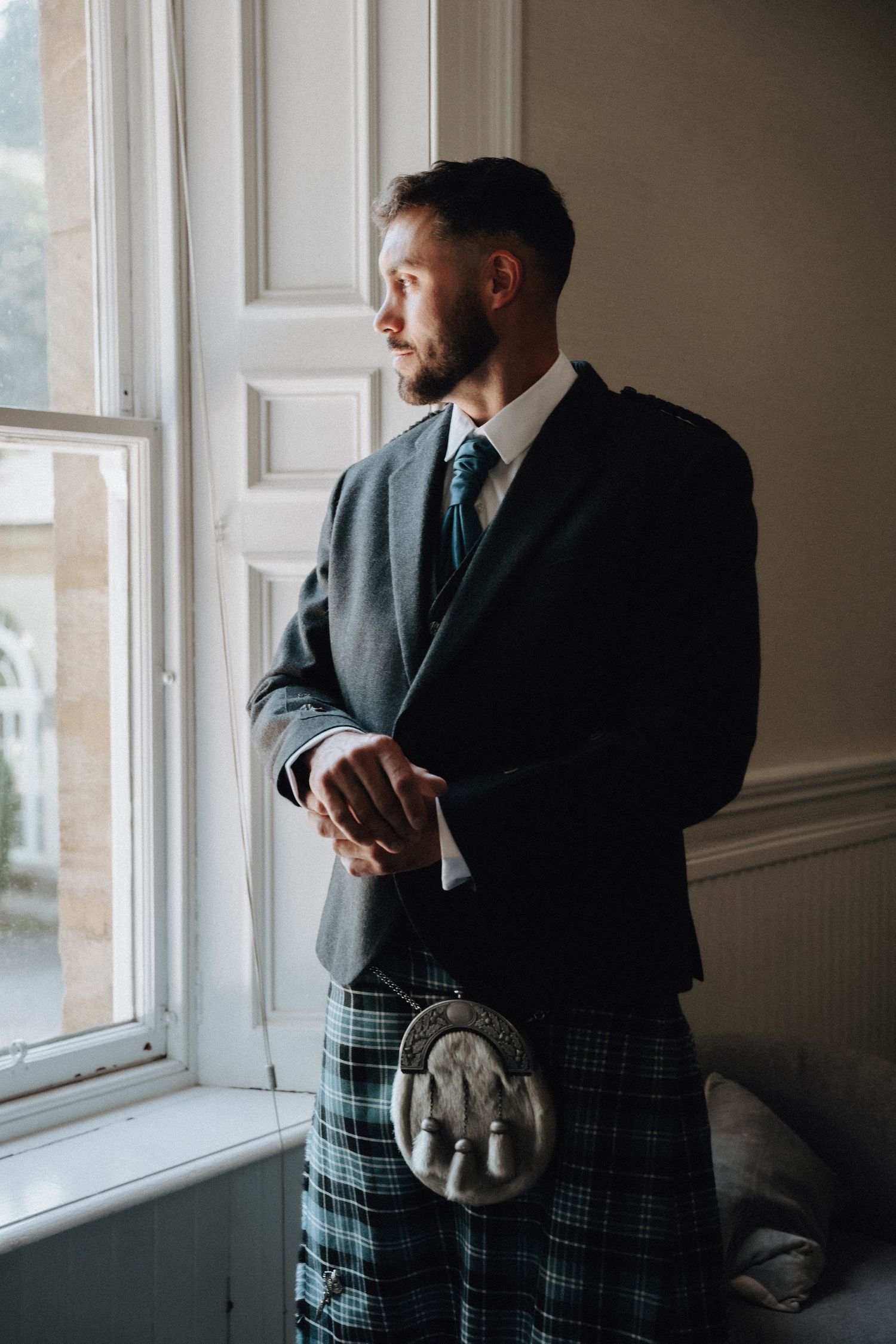 Person in formal suit and kilt stands pensively by a bright window in an elegant room.