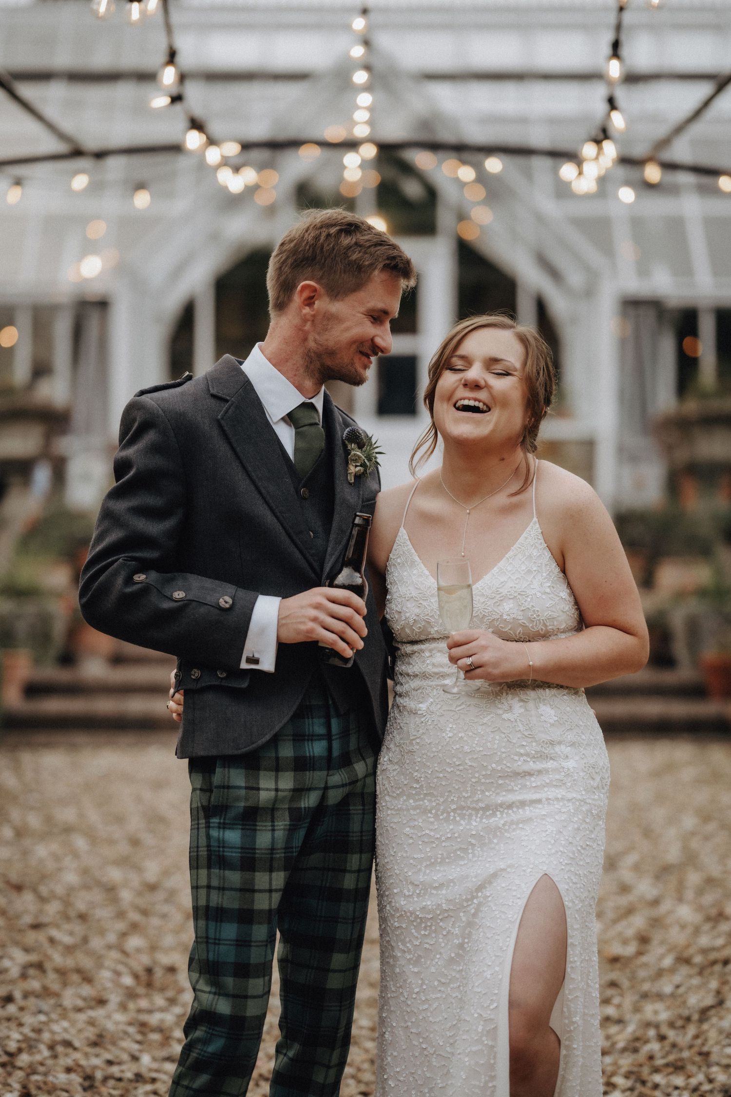 Couple shares a laugh in greenhouse setting with string lights overhead.