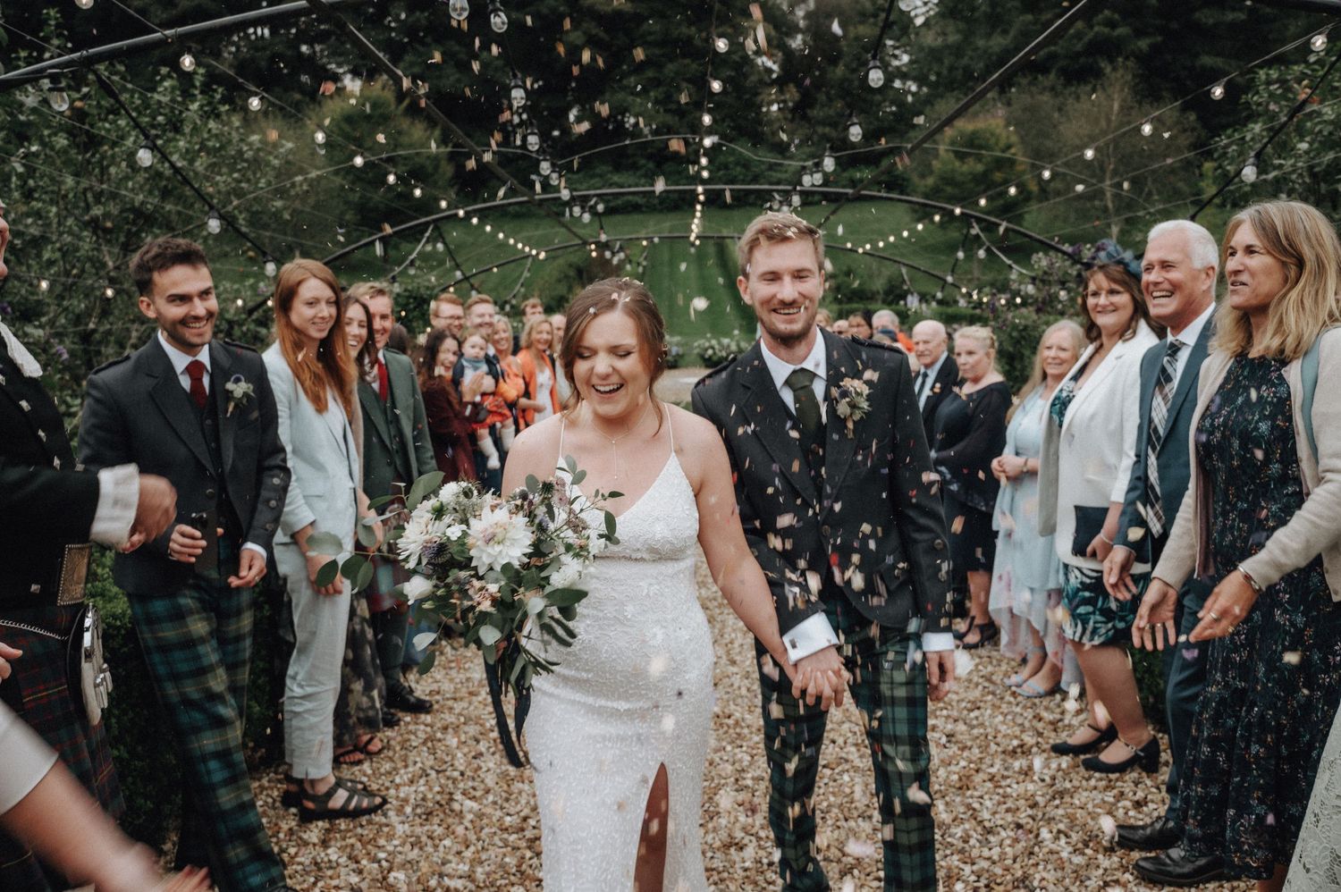 Newlyweds walk through shower of confetti with guests cheering along path.