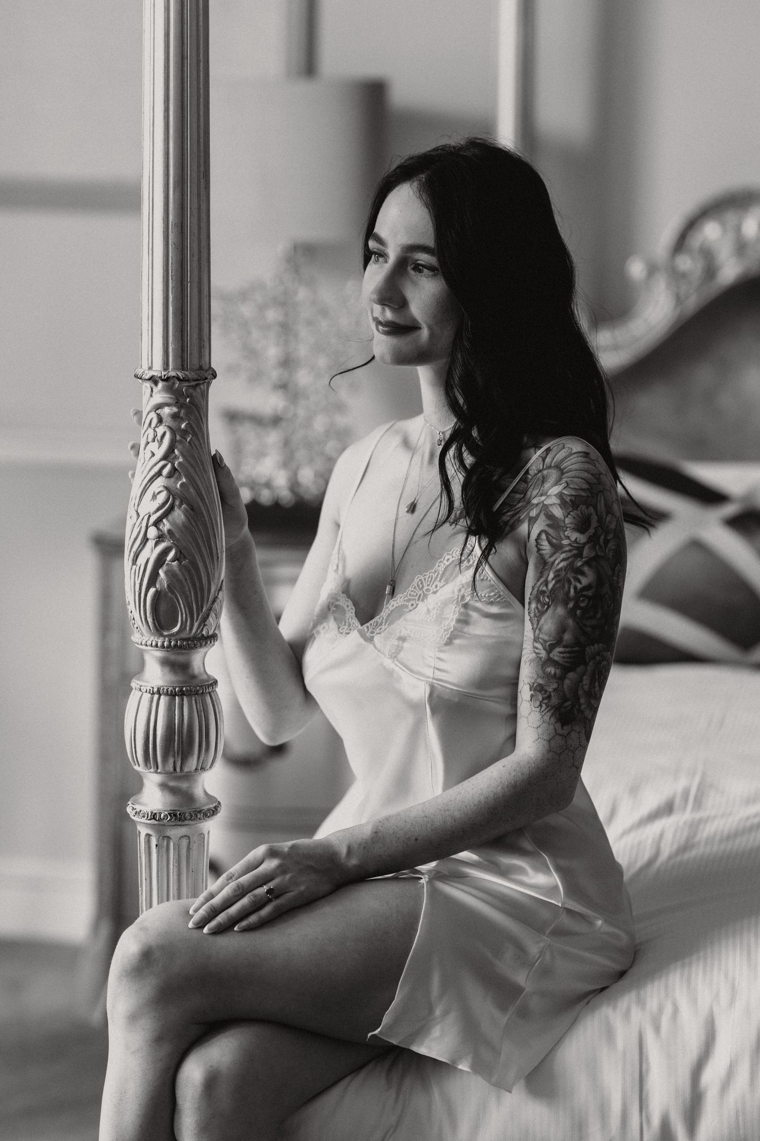 Woman in white silk slip dress sitting on bed beside ornate bedpost in black and white photo.