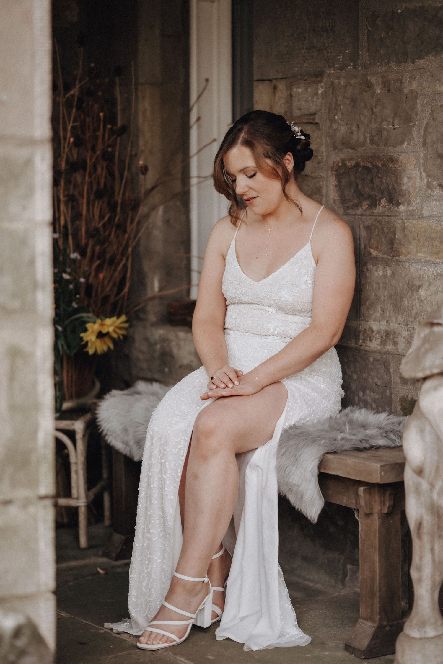 A person in a white lace dress sits on a rustic bench against a stone wall.