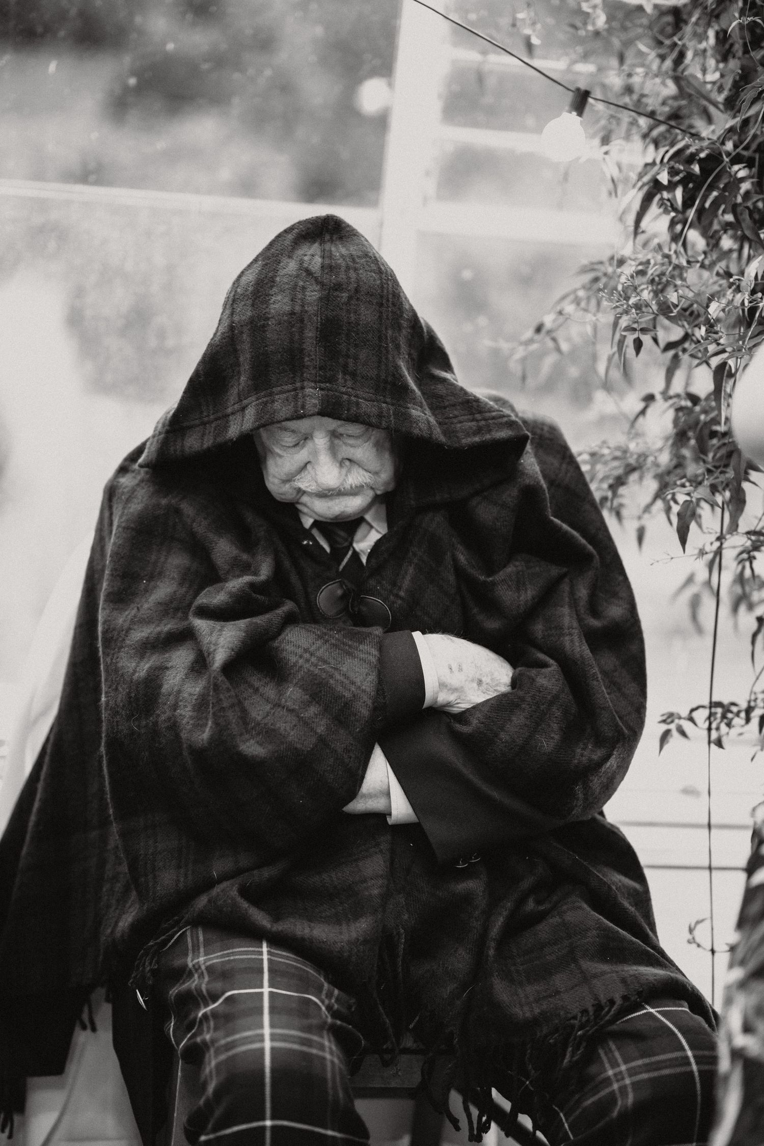 Person wrapped in plaid cloak sleeping in chair at outdoor event.
