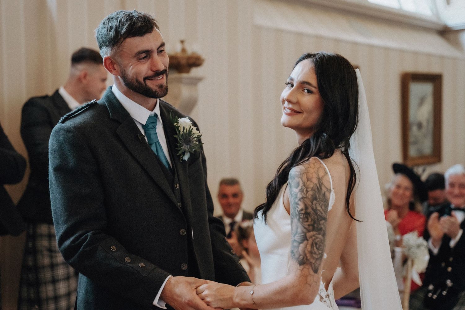 Couple holding hands at the altar with bride in white dress and groom in traditional Scottish attire.